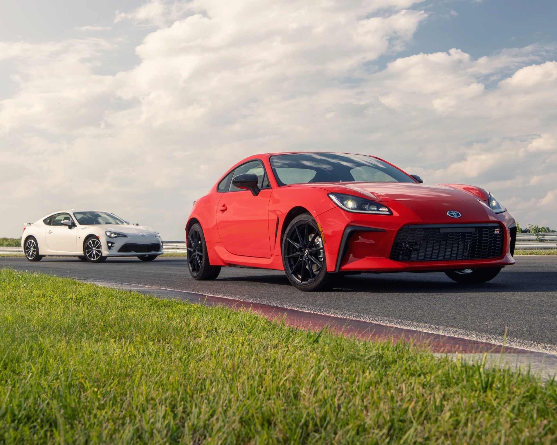 Two sporty Toyota GR cars on a racetrack, with a red model in the foreground and a white model behind it, both featuring sleek designs and black wheels under a partly cloudy sky.