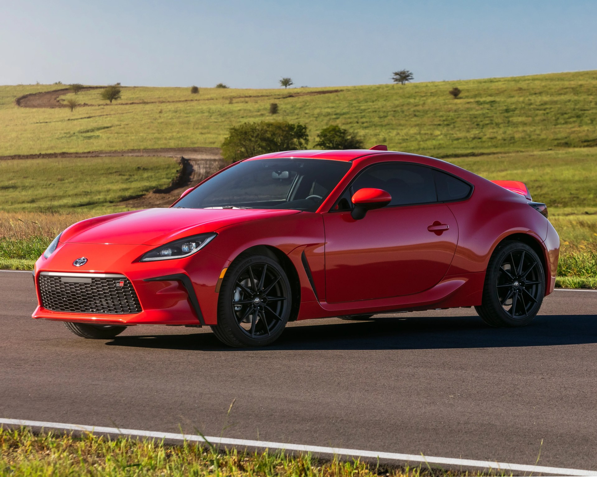 Red two-door sports coupe with black wheels parked on a paved road, set against a backdrop of green grassy hills and a clear blue sky. The car features a sleek, aerodynamic design with a prominent front grille and sharp headlights.