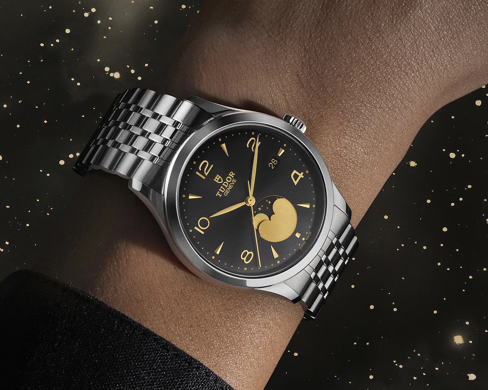 Silver Tudor wristwatch with a black dial featuring gold hour markers and hands, a date window at 3 o'clock, and a gold moon phase indicator at 6 o'clock, worn on a wrist against a dark background with gold speckles.