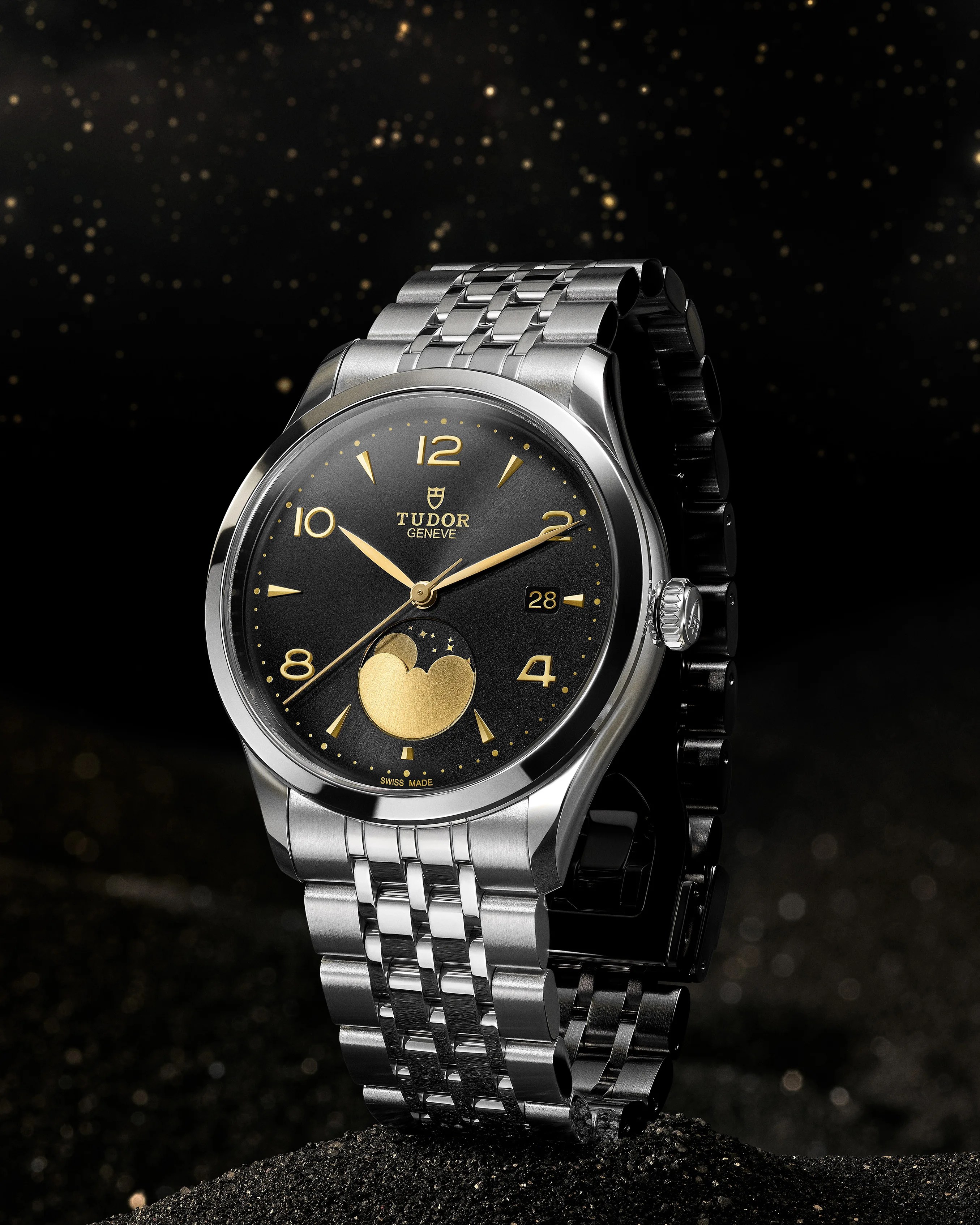 Silver Tudor wristwatch with a black dial featuring gold hour markers and hands, a moon phase indicator at 6 o'clock, and a date window at 3 o'clock. The watch has a polished stainless steel case and a multi-link metal bracelet, set against a dark, starry background.