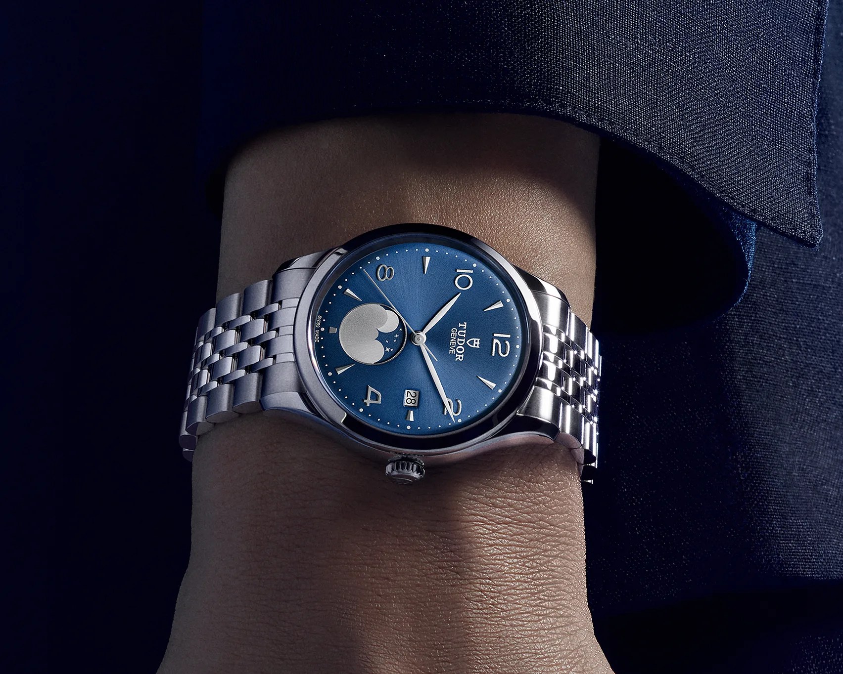 Silver Tudor wristwatch with a blue dial featuring silver hour markers, a moon phase indicator at 6 o'clock, and a date window at 3 o'clock, worn on a wrist with a silver metal bracelet.