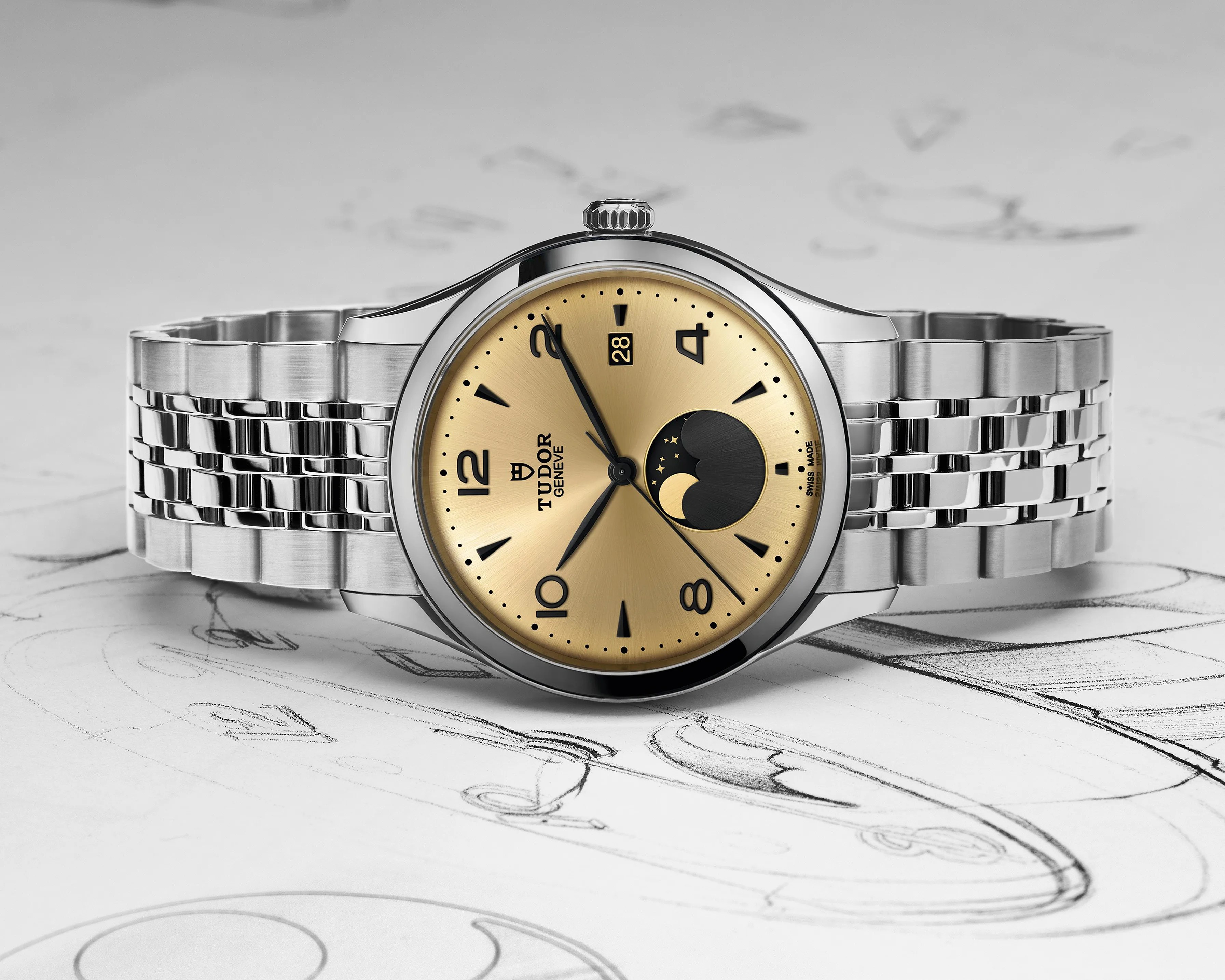 Silver Tudor wristwatch with a gold dial featuring black Arabic numerals at 2, 4, 8, 10, and 12 o'clock, black hour markers, and a moon phase indicator at 6 o'clock. The watch has a date window at 12 o'clock and a stainless steel bracelet with polished and brushed links. The background shows sketches of watch designs.