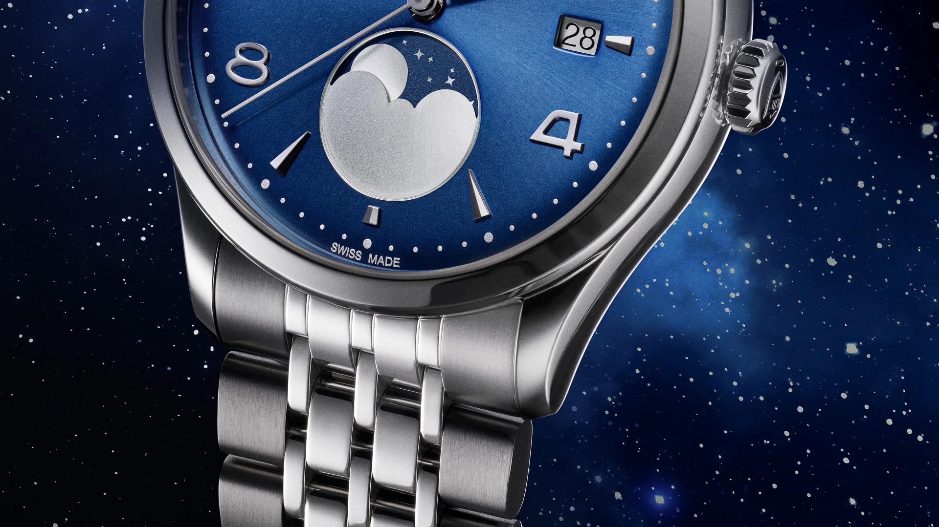 Close-up of a luxury wristwatch with a blue dial featuring a moon phase indicator, silver hour markers, and a date window showing the 28th. The watch has a polished stainless steel case and a linked metal bracelet. The background resembles a starry night sky.