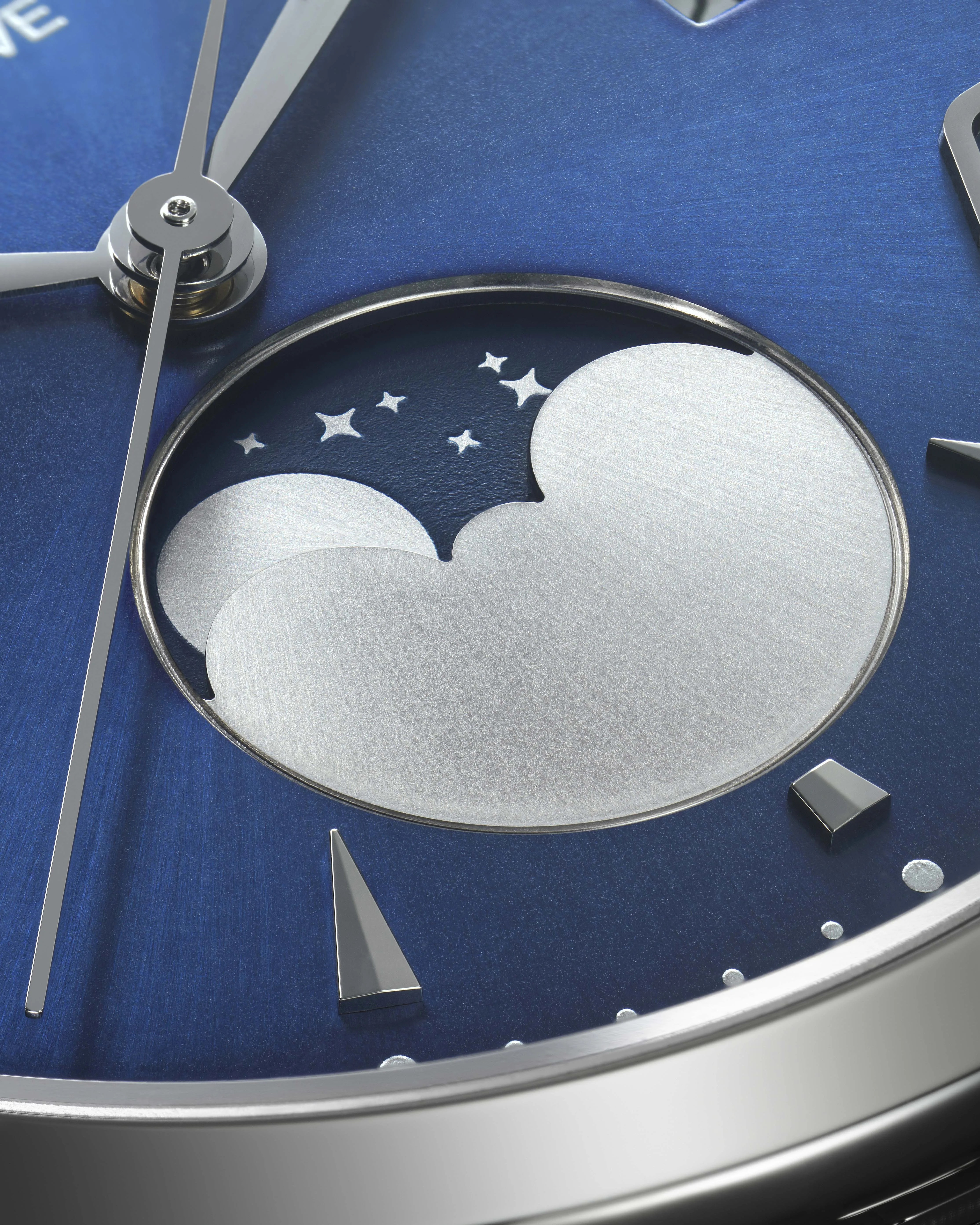 Close-up of a blue watch dial featuring a moon phase complication. The moon phase display shows a silver moon and stars against a dark blue background, with polished silver watch hands and triangular hour markers nearby. The watch case is metallic with a brushed finish.
