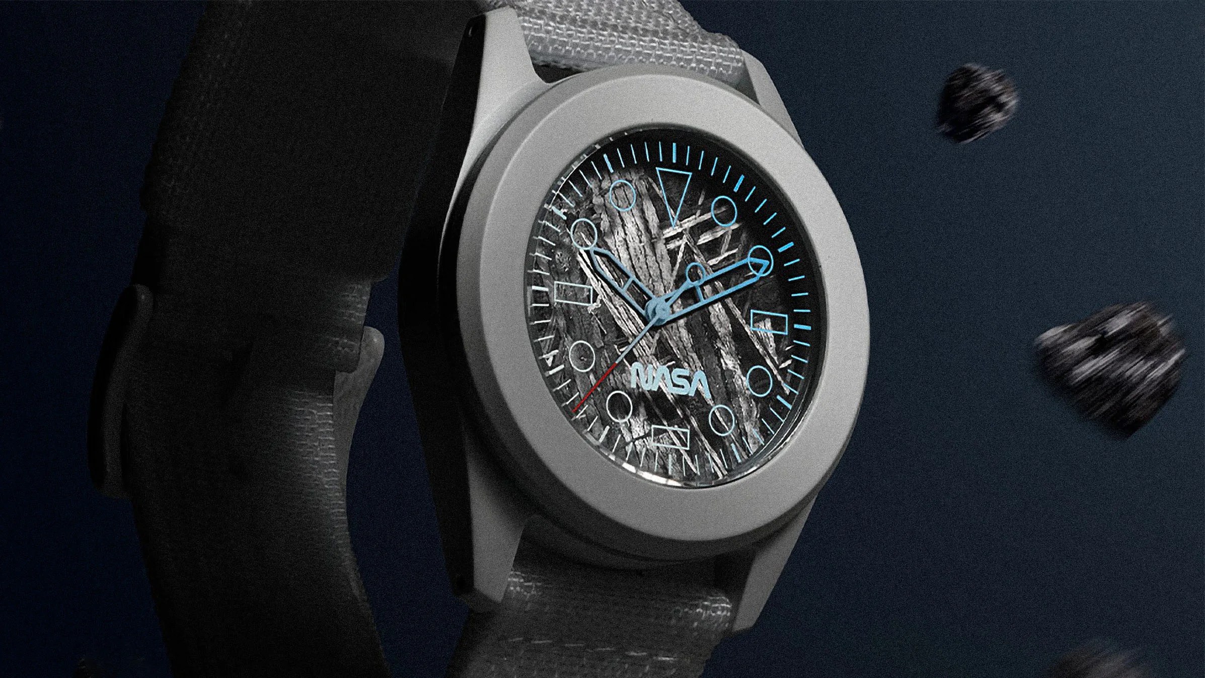 A close-up of a wristwatch with a gray strap and a round silver bezel. The watch face has a textured, rocky background with light blue hour markers and hands, and a red second hand. The word "NASA" is printed in light blue near the bottom center of the watch face. The background is dark with small floating rock-like objects.