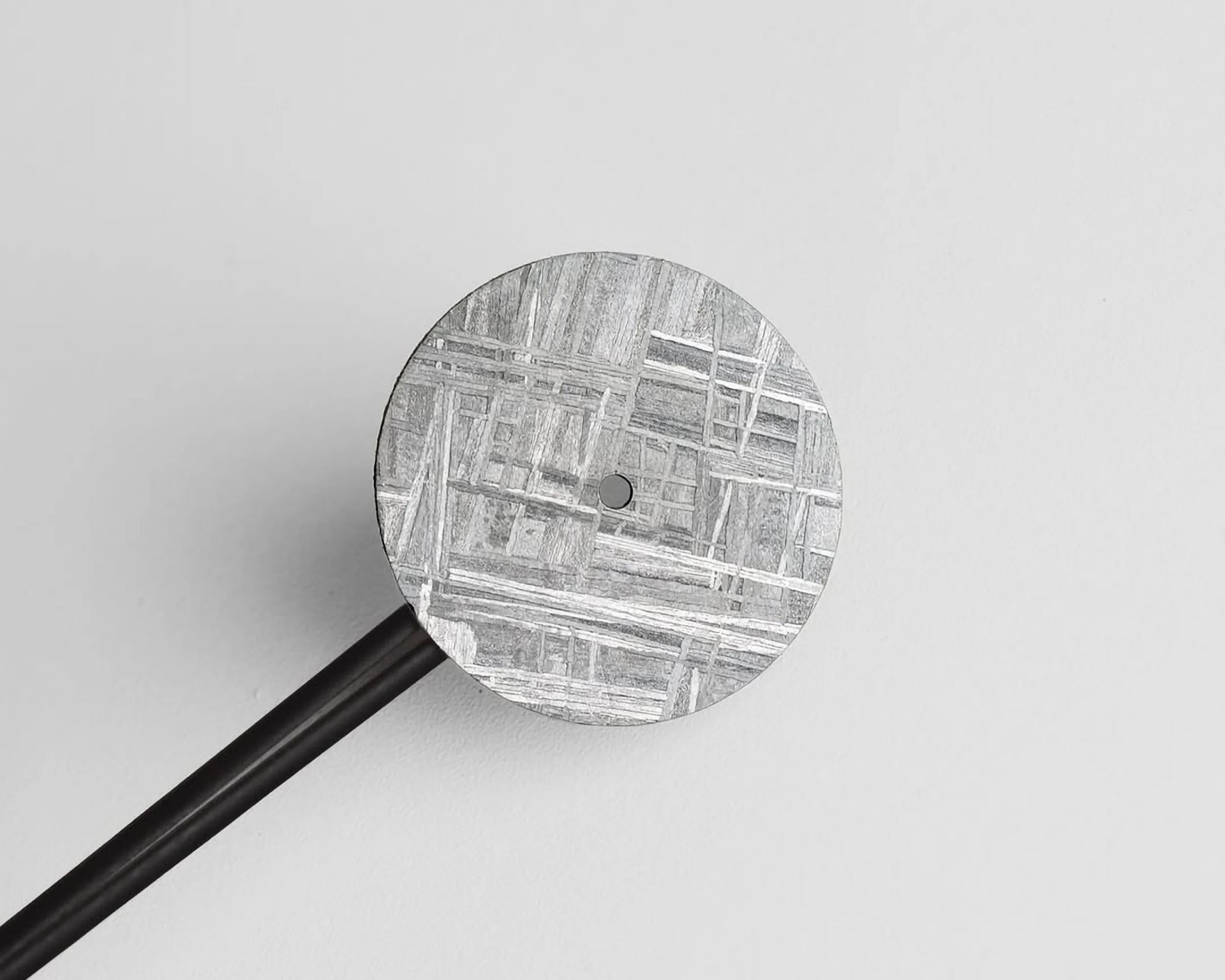 A close-up of a round metallic object with a textured, crisscross pattern on its surface, attached to a black rod or handle, set against a plain light gray background.