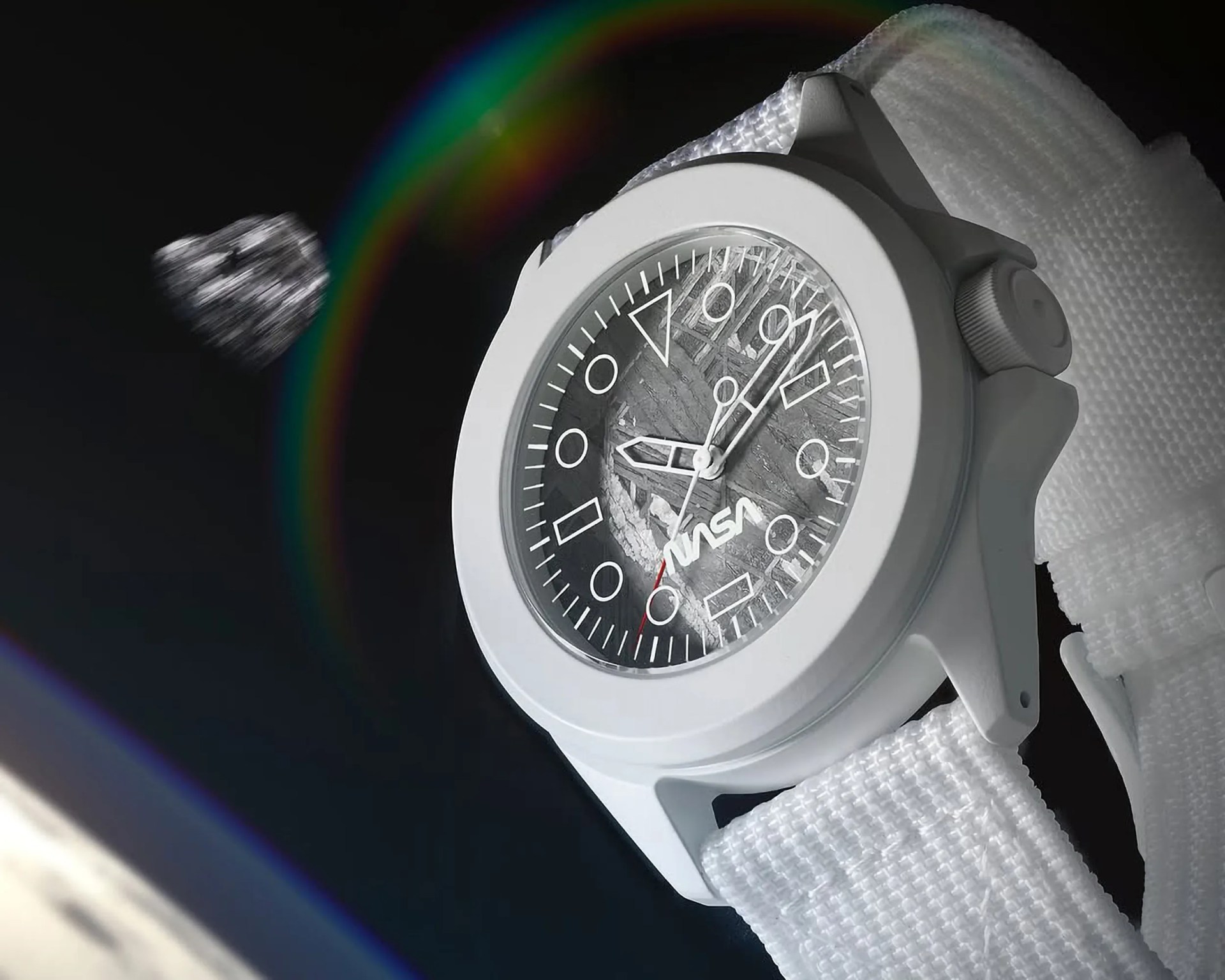 A close-up of a white NASA-themed wristwatch with a textured dial resembling a lunar surface. The watch has white geometric hour markers and hands, a red second hand, and a white fabric strap. A faint rainbow-like lens flare appears in the background against a dark backdrop.