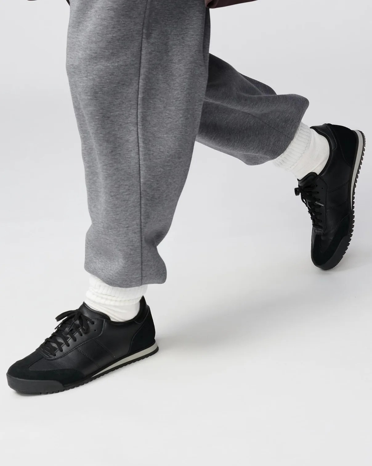 Black low-top sneakers with a combination of smooth and suede textures, featuring black laces and a white midsole with a black outsole. The shoes are worn with white socks and gray sweatpants.