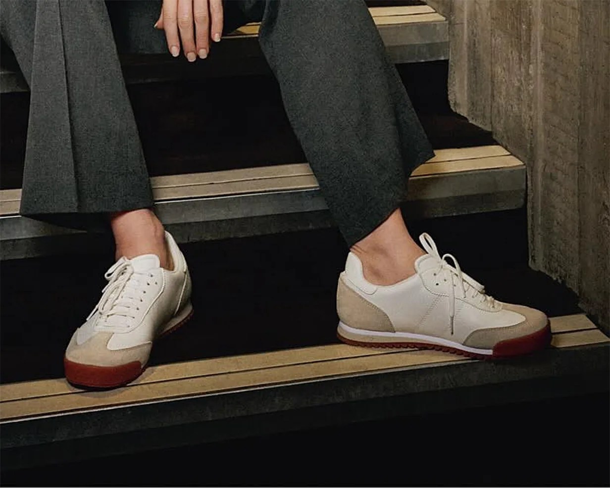White sneakers with beige suede accents and red rubber soles, worn with dark gray pants, shown on a person sitting on stairs with a concrete wall background.