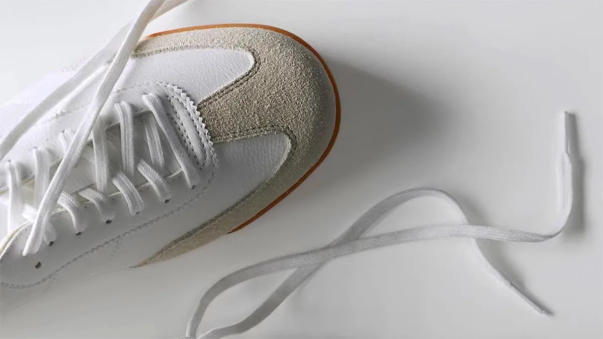 White sneaker with beige suede accents on the toe and a brown sole edge, featuring white laces, one of which is untied and extended on a white surface.