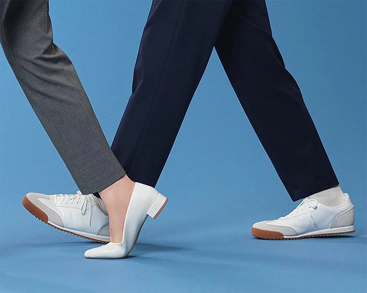 Two pairs of legs are shown walking on a blue background. One person is wearing gray pants with white and beige sneakers that have brown soles. The other person is wearing dark navy pants with white heeled shoes.