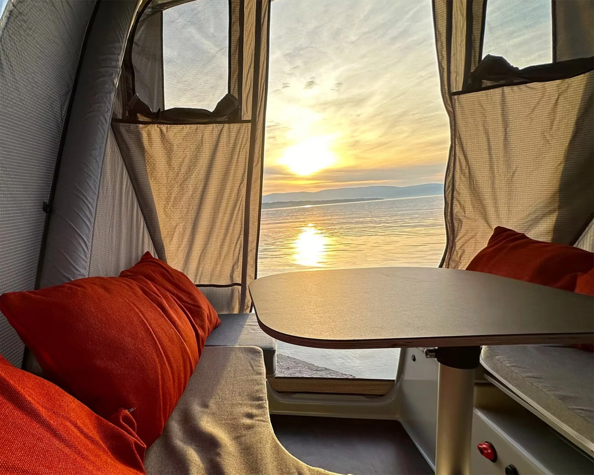 Interior seating area of a tent or camper with a table in the center, cushioned benches on either side, and red pillows. The tent's open flap reveals a view of a calm body of water with the sun setting or rising over distant hills.
