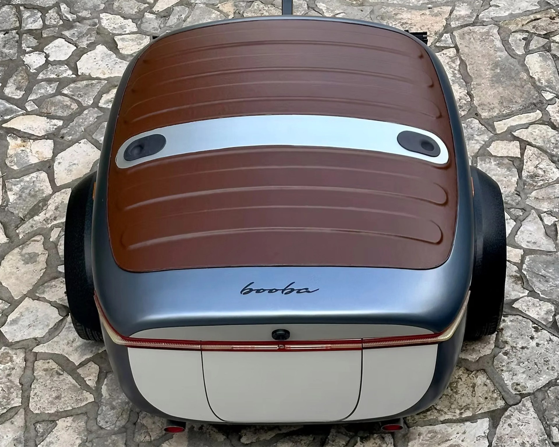 A top-down view of a small, compact car with a brown roof featuring a silver strip with two black circular elements. The car's rear is silver with the word "booba" written in a cursive style. The car is parked on a stone-paved surface.