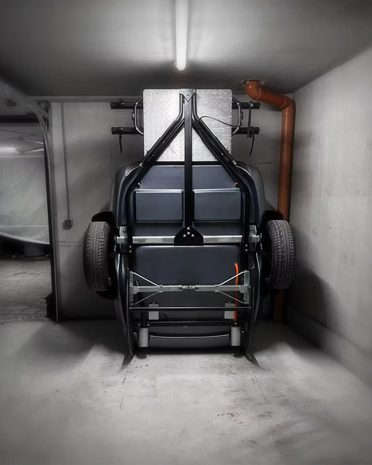 A compact, black, three-wheeled vehicle is parked vertically against a wall in a concrete garage. The vehicle's underside, including its frame, tires, and mechanical components, is visible. A fluorescent light fixture is mounted on the ceiling above the vehicle, and an orange pipe runs vertically along the right wall. The garage has a utilitarian, industrial appearance with plain concrete walls and floor.