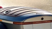 Rear view of a blue and white boat with a ribbed black cover and a horizontal red tail light strip. The boat is on water, and there is a small, partially visible red object on the left side.