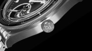 Close-up of a luxury wristwatch crown and part of the bezel and case, featuring a brushed metal finish and a detailed engraved logo on the crown, with a black background.