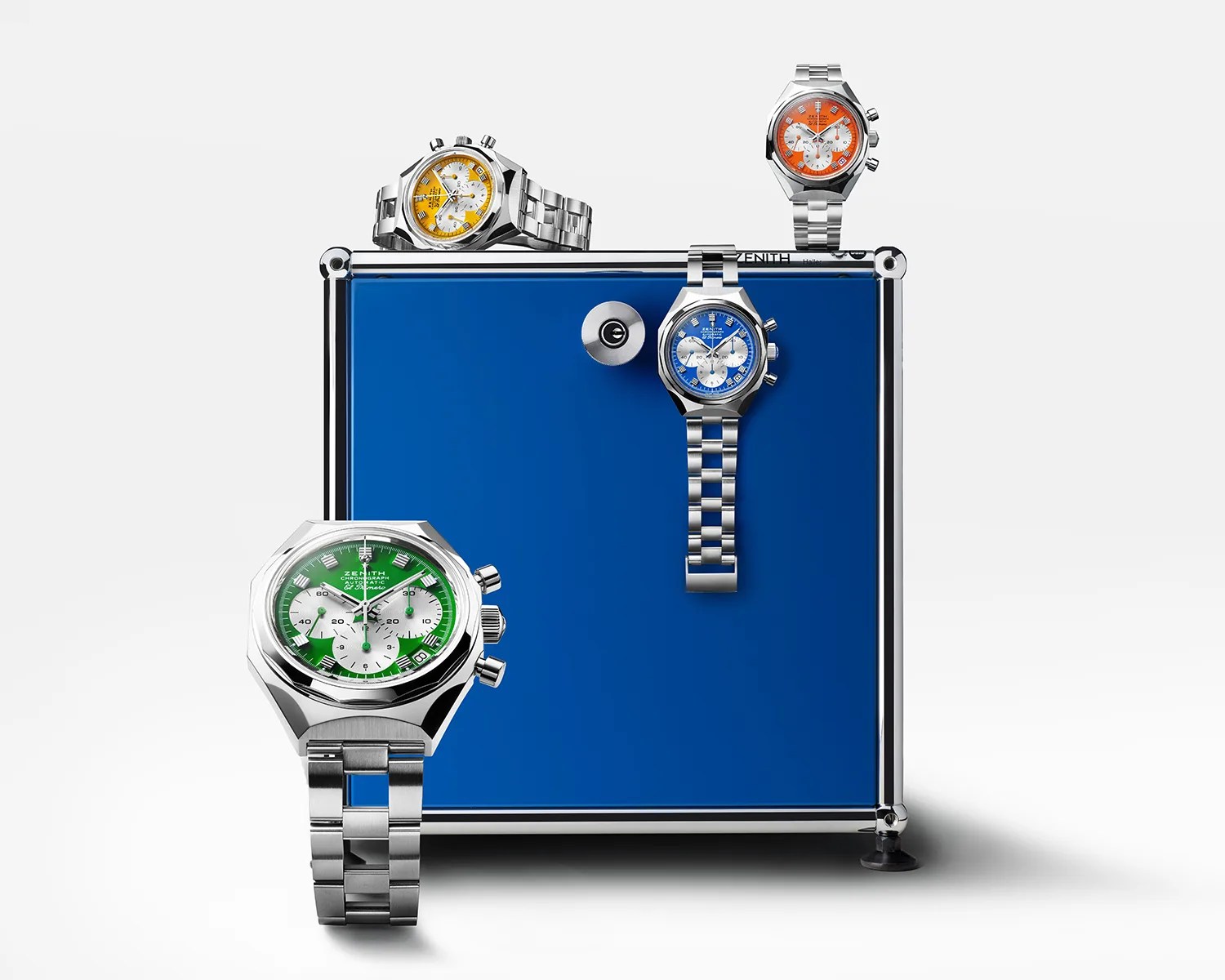 Four stainless steel wristwatches with colorful dials—green, blue, yellow, and orange—are displayed around a blue and silver metal box. Each watch features a chronograph with three subdials and a date window, and they have matching stainless steel link bracelets. The watches are arranged with one green watch in front, a blue watch hanging on the box, a yellow watch resting on top, and an orange watch standing upright on the box.