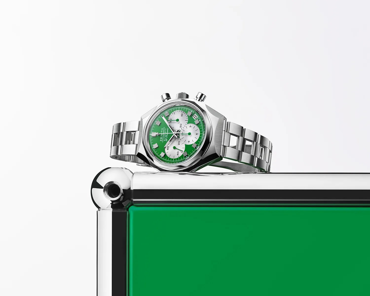 Silver wristwatch with a green dial and three white subdials, featuring a stainless steel bracelet, placed on the corner of a green and chrome metallic surface. The watch has two pushers and a crown on the right side of the case.