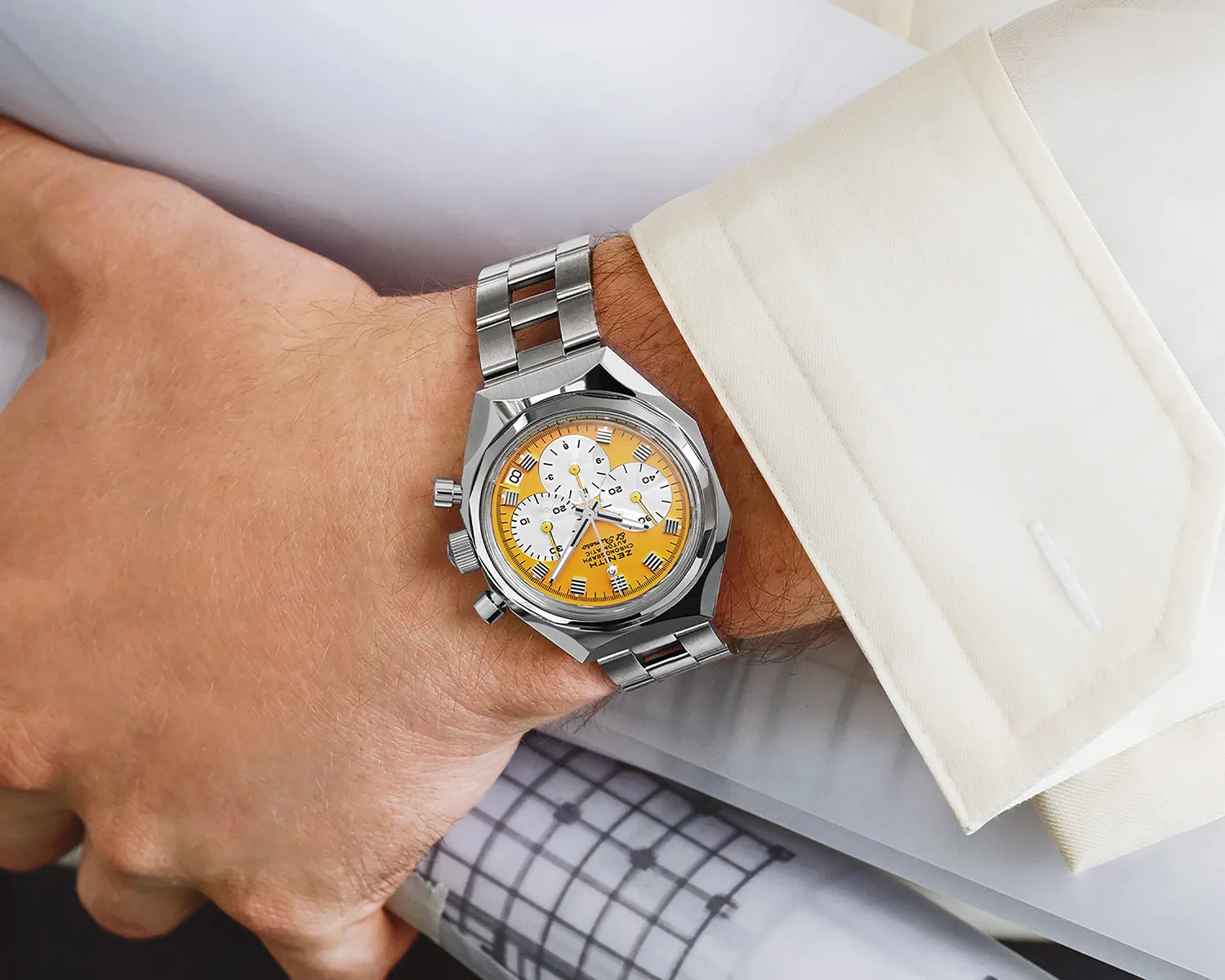 Silver metal wristwatch with a yellow dial and three white subdials, worn on a wrist with a white dress shirt cuff. The watch has a polished, angular case and a linked metal bracelet. The dial features hour markers and chronograph functions.