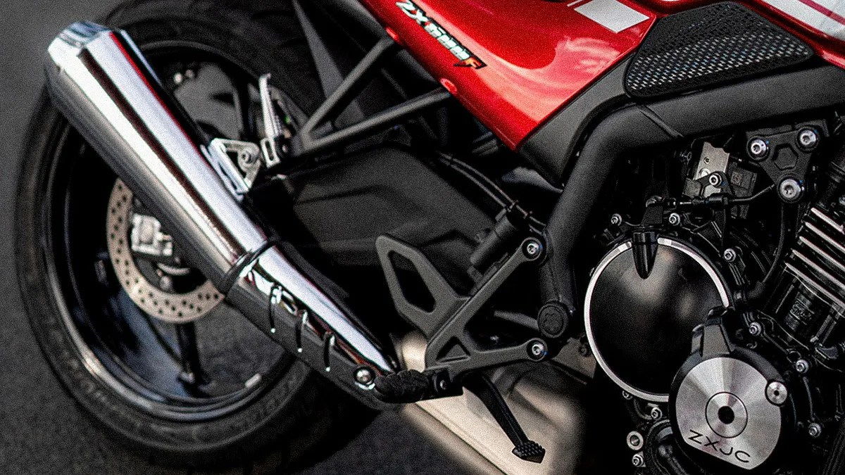 Close-up of a red motorcycle's rear section showing the exhaust pipe, rear wheel with disc brake, footpeg, and part of the engine with "ZXJC" engraved on a circular metal cover. The frame and various mechanical components are also visible.