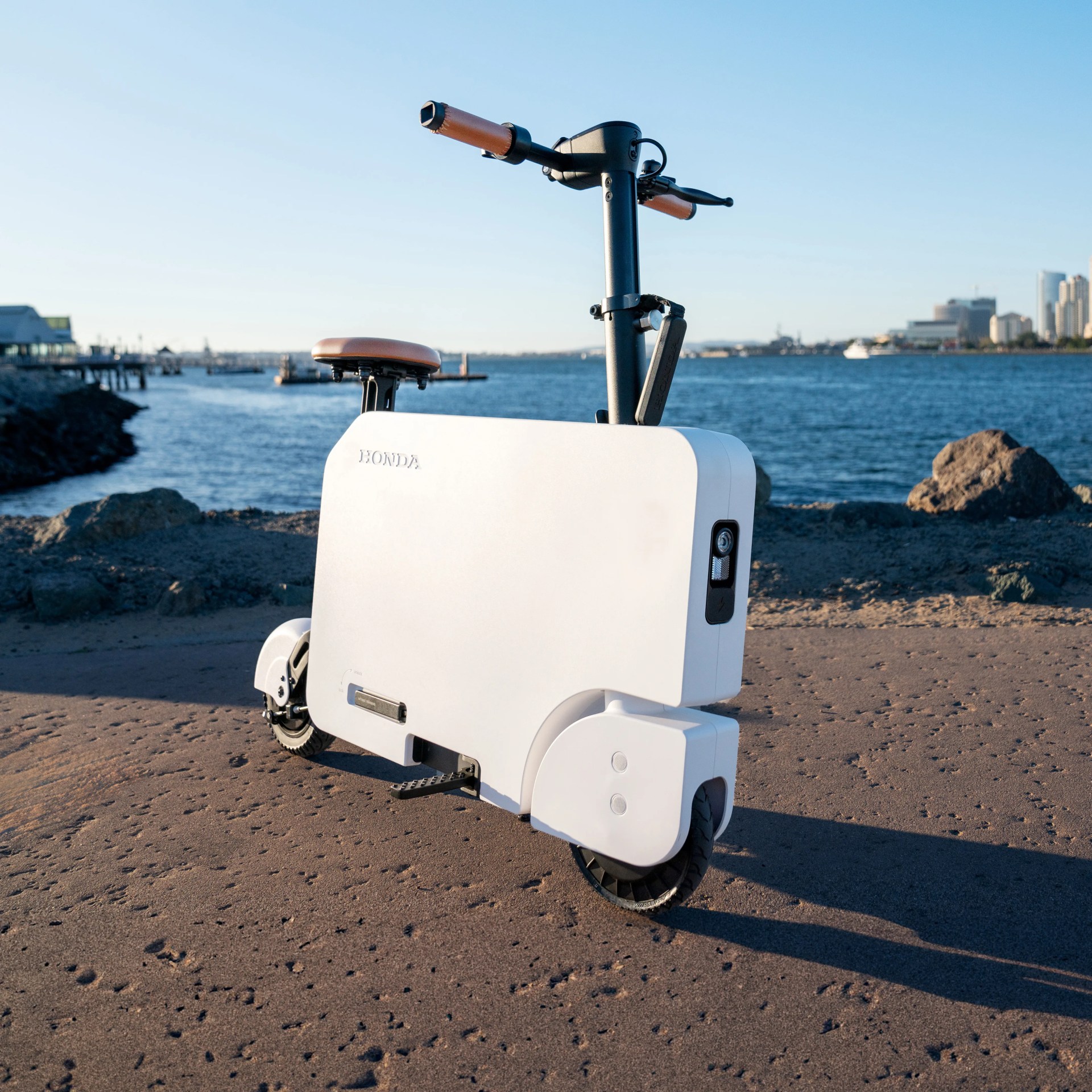 White compact Honda electric scooter with brown seat and handle grips on a waterfront path.
