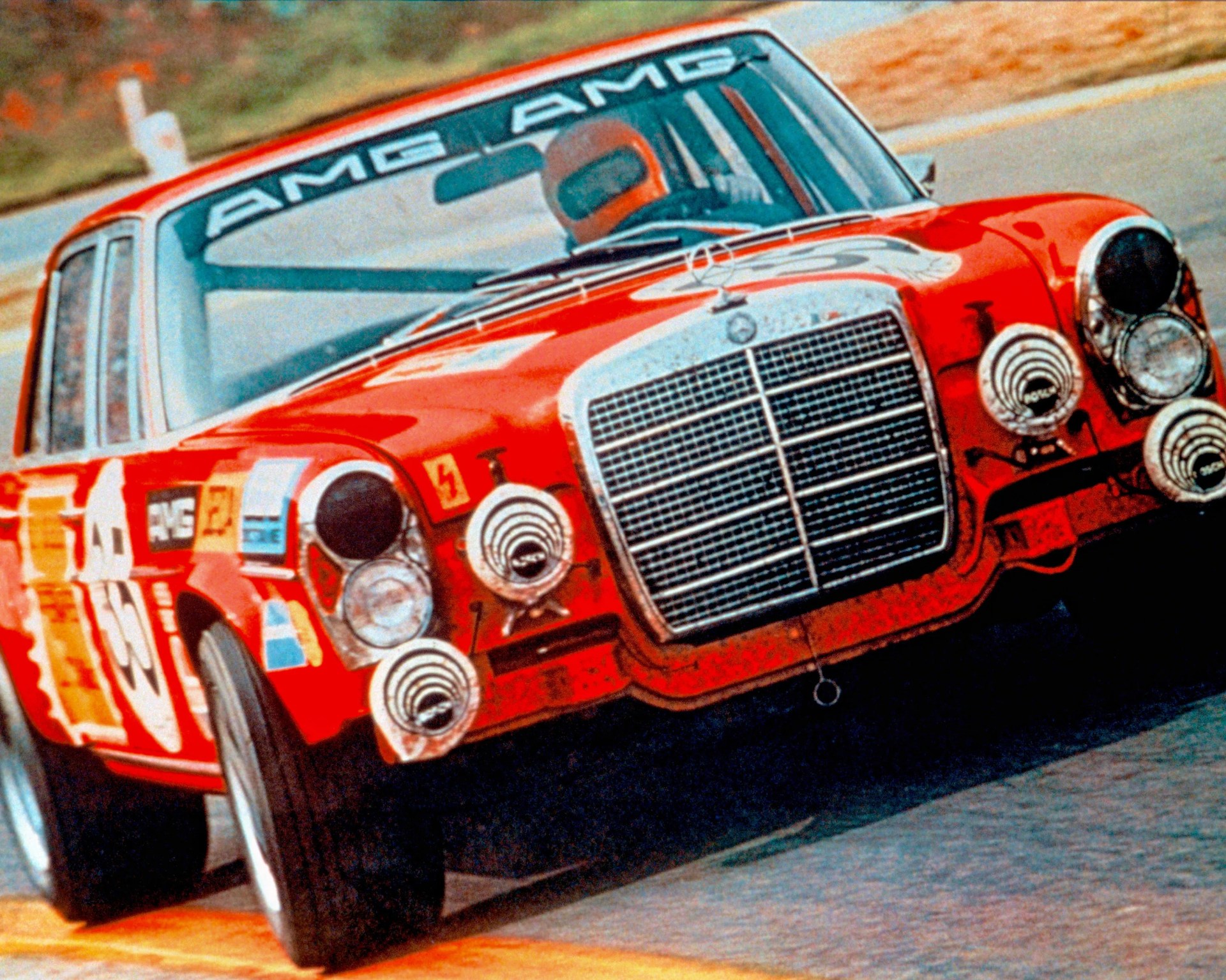 Red vintage Mercedes-Benz AMG race car with multiple front spotlights on a racetrack.