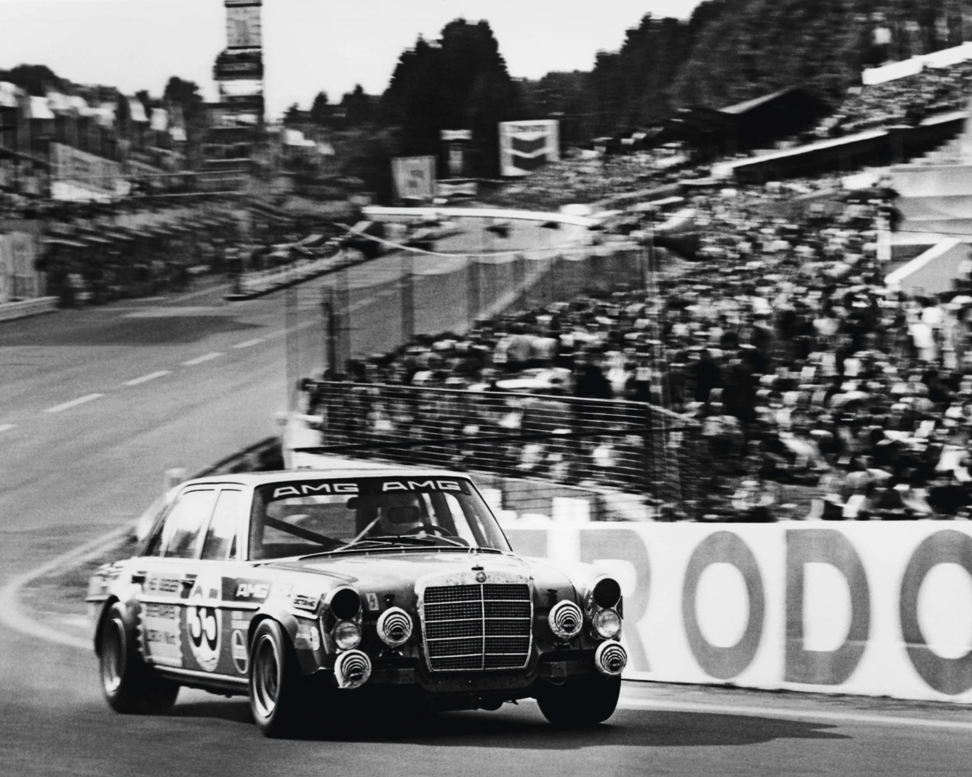 Black and white photo of a vintage Mercedes-Benz AMG race car number 55 on a racetrack with a crowd in the background.