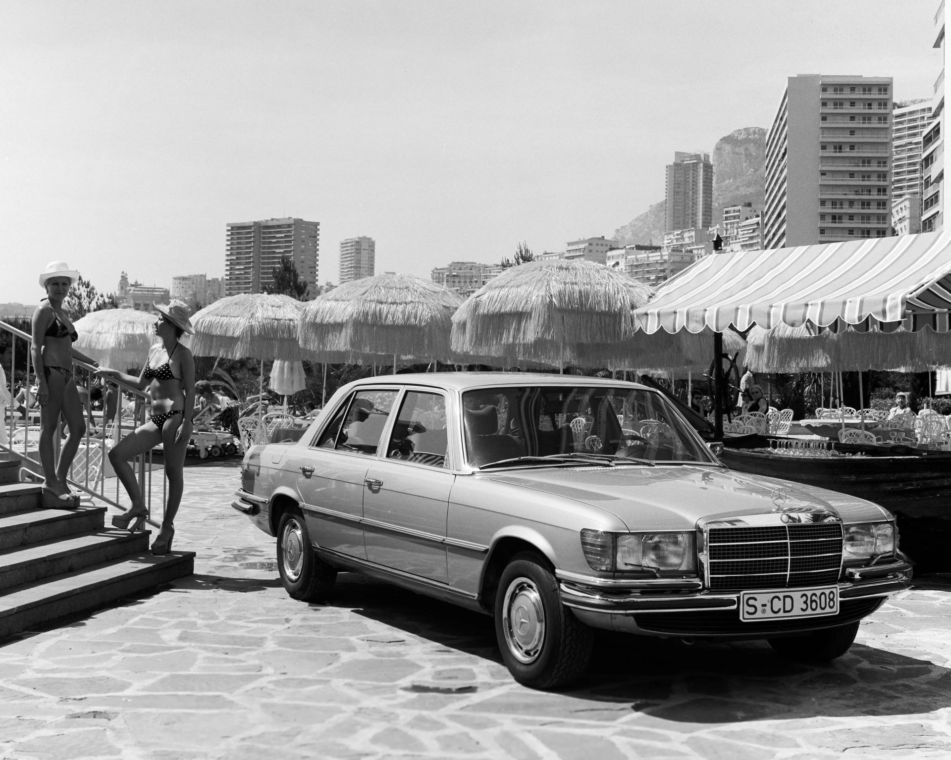 Classic Mercedes-Benz sedan parked on a stone patio near straw umbrellas and two women in bikinis on stairs.