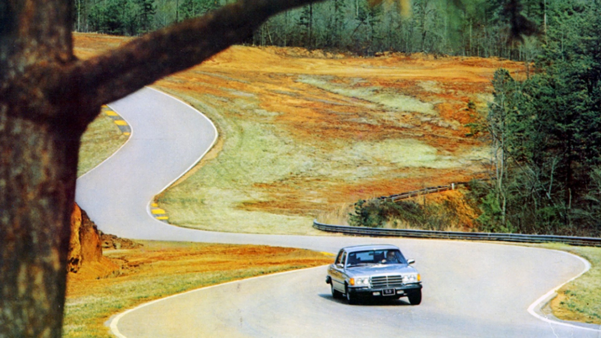 Silver sedan car driving on a winding paved road surrounded by autumn-colored grass and trees.