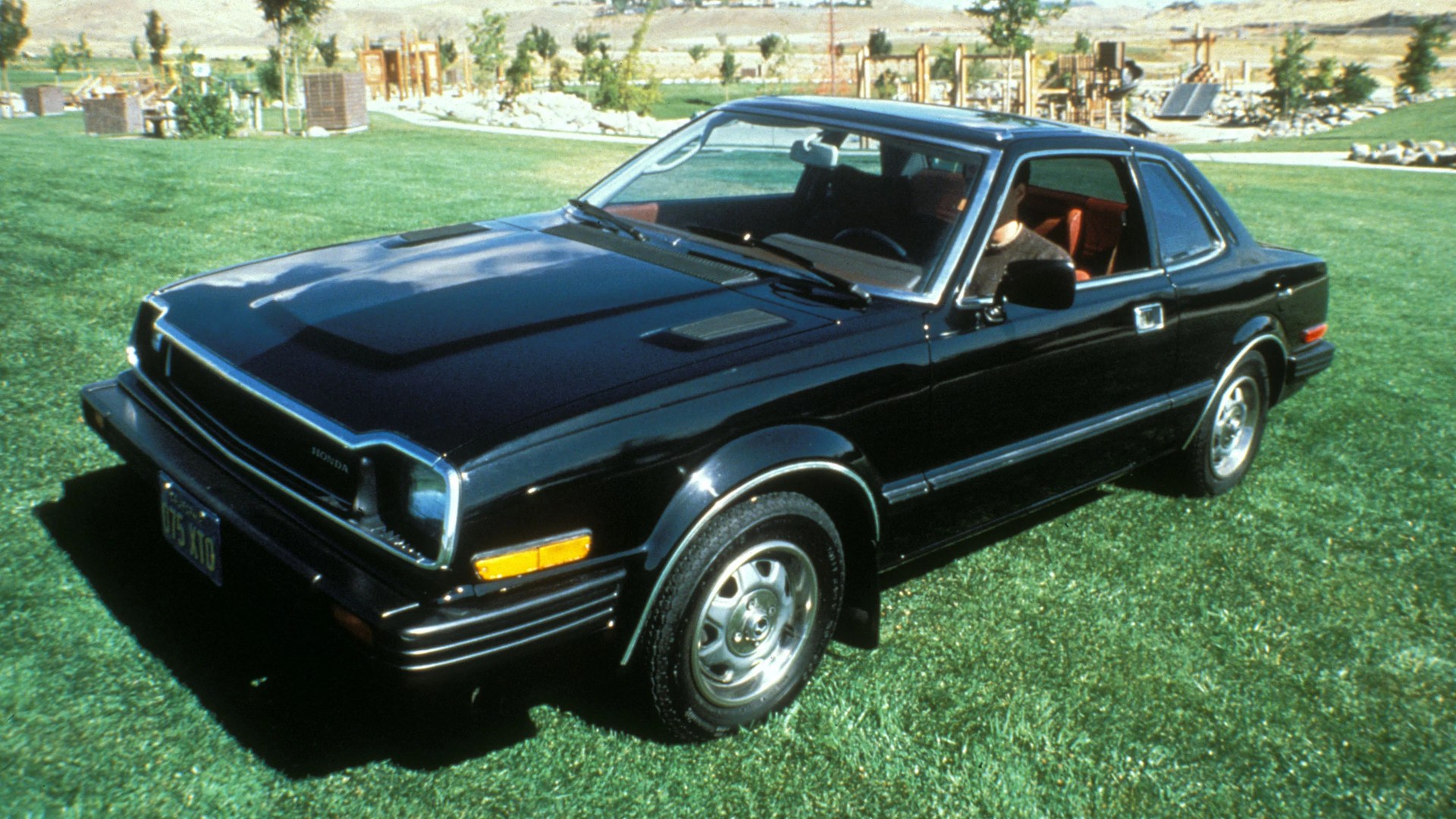 Black two-door vintage Honda coupe parked on green grass with a person inside.