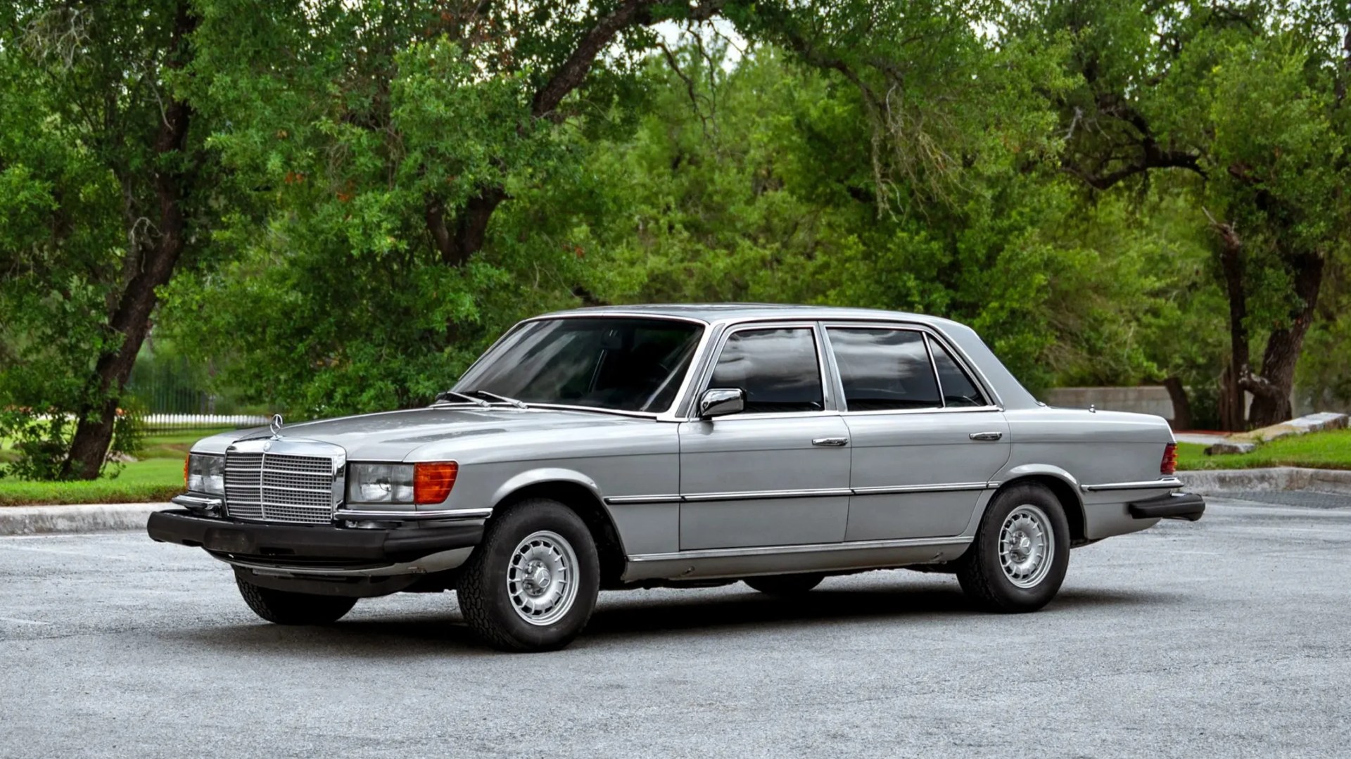 Silver vintage Mercedes-Benz sedan parked on a paved area with green trees in the background.