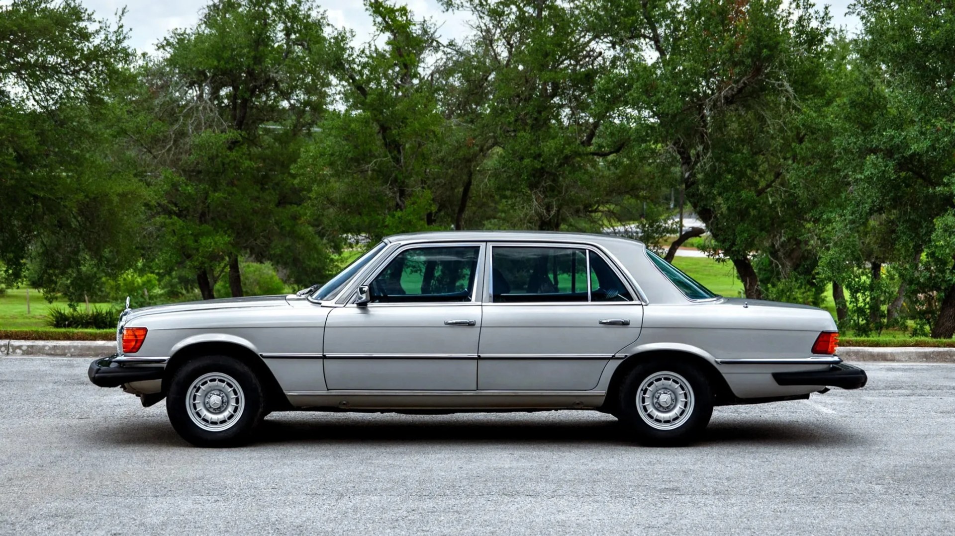 Silver vintage Mercedes-Benz sedan parked on a paved surface with green trees in the background.