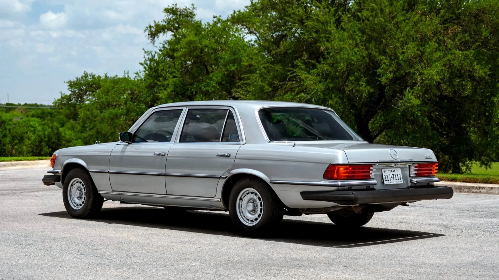 Silver vintage Mercedes-Benz sedan parked on a paved surface with green trees in the background.