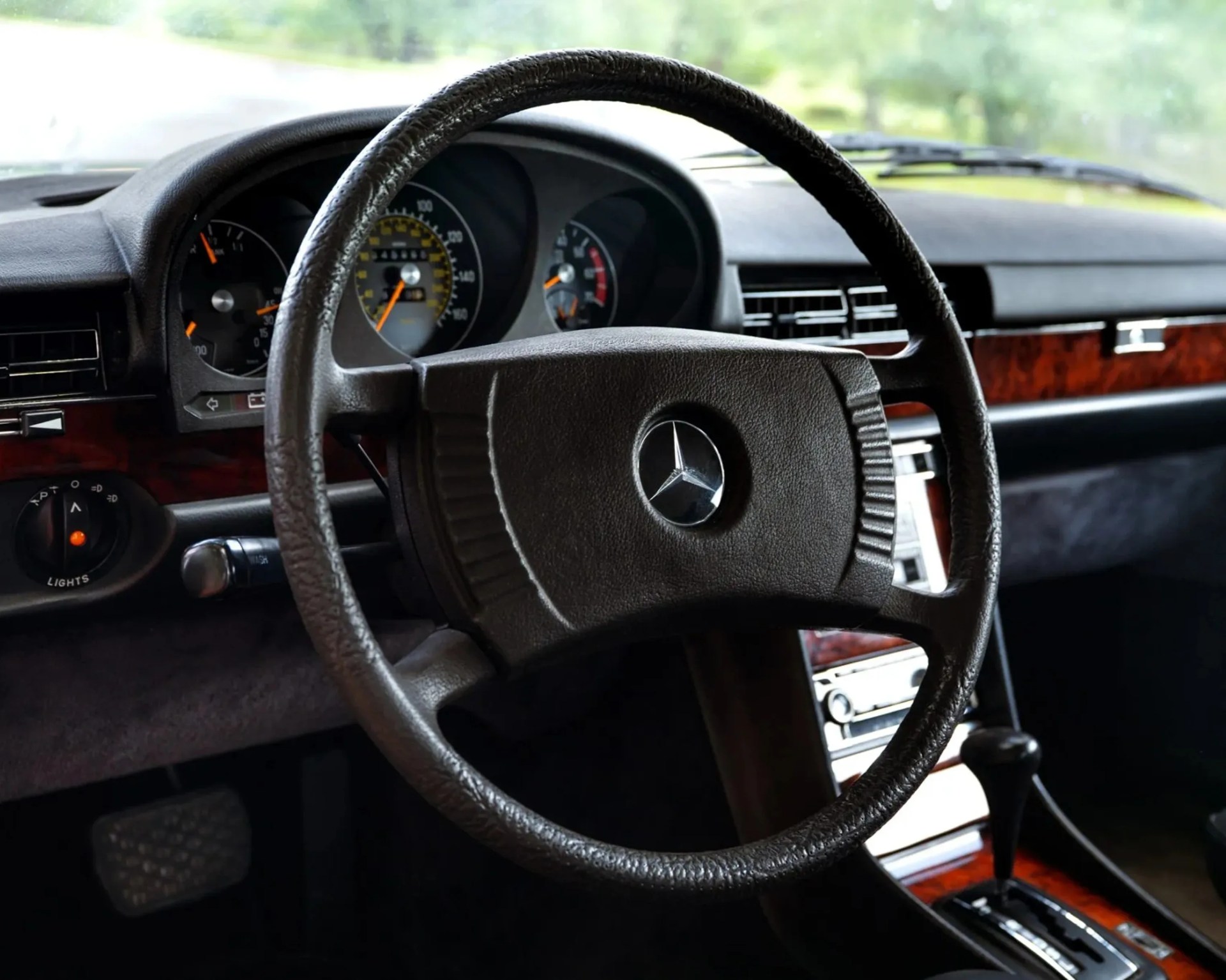 Black leather-wrapped steering wheel with Mercedes-Benz logo inside a car dashboard with wood trim and automatic gear shift.