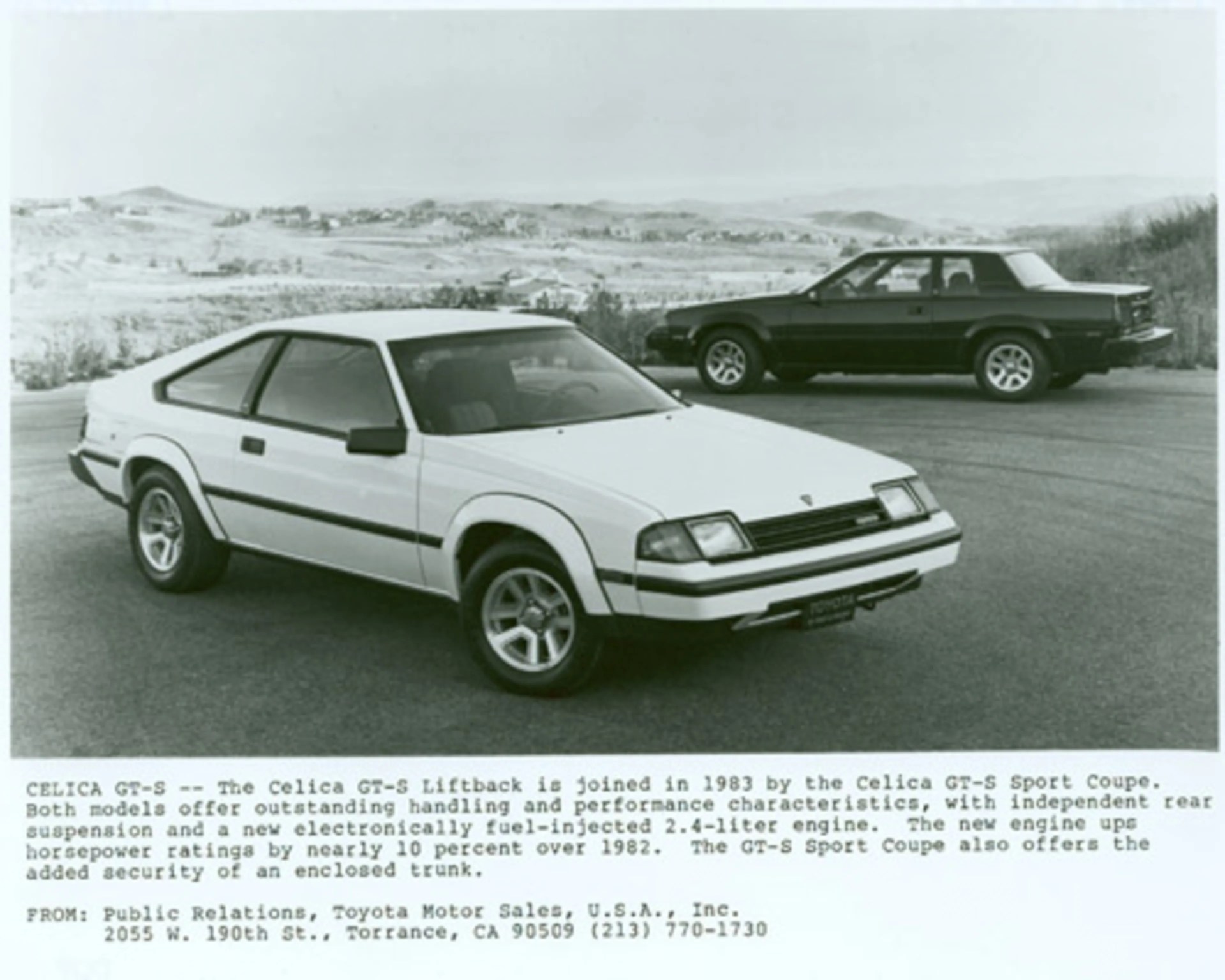 Two 1983 Toyota Celica GT-S cars, one white liftback in the foreground and one black sport coupe in the background, parked on a paved area with hills in the distance.