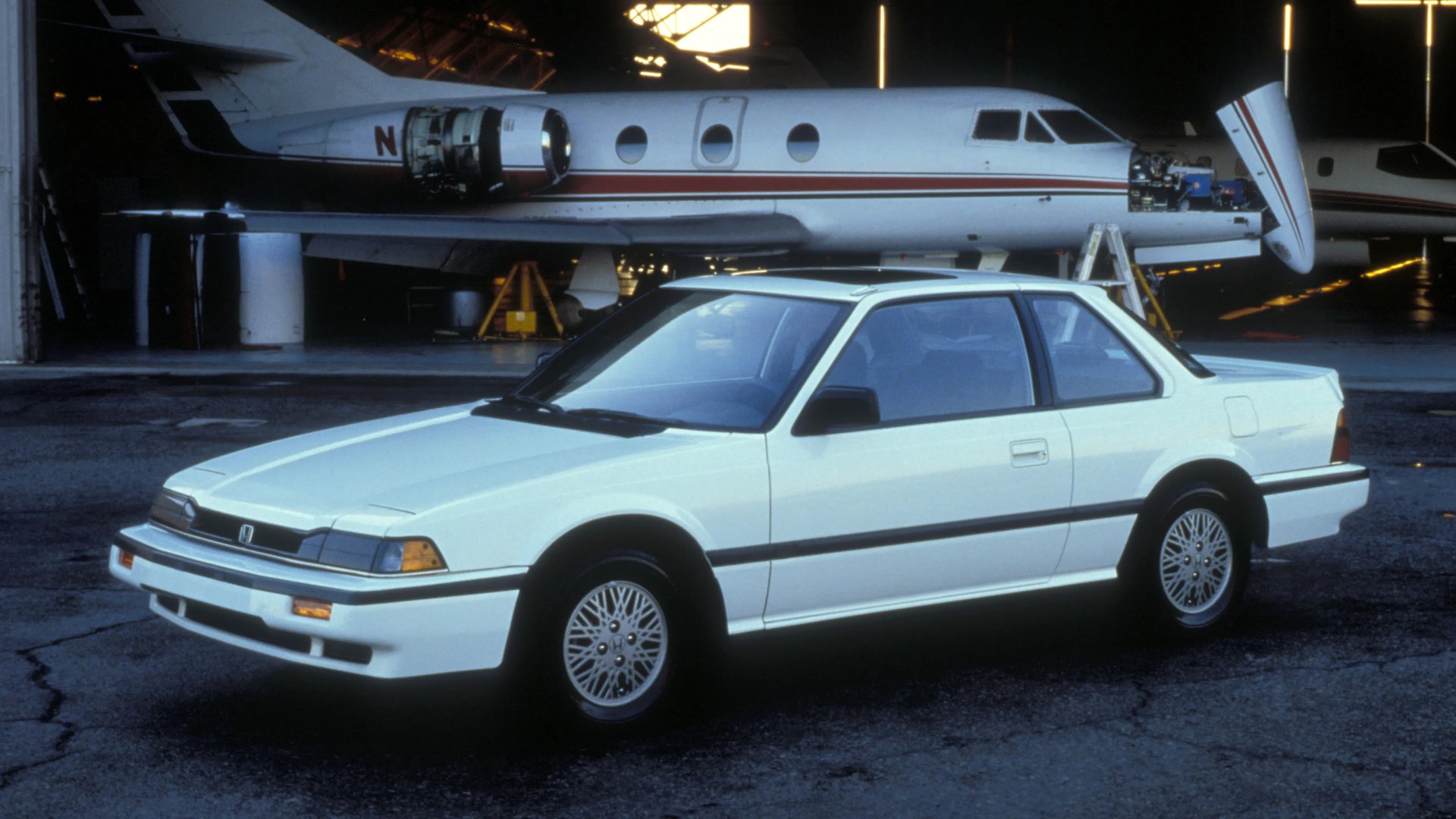 White two-door Honda coupe parked on tarmac in front of a private jet with open engine panels.