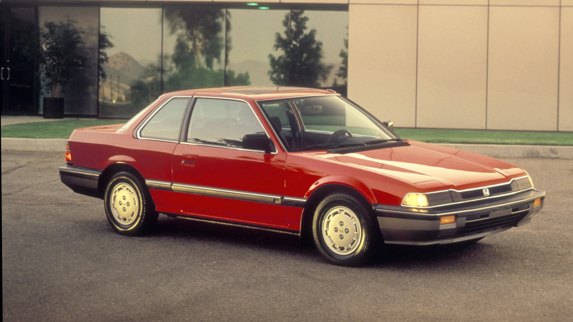 Red two-door Honda coupe with silver trim and alloy wheels parked on asphalt near a building with reflective windows.