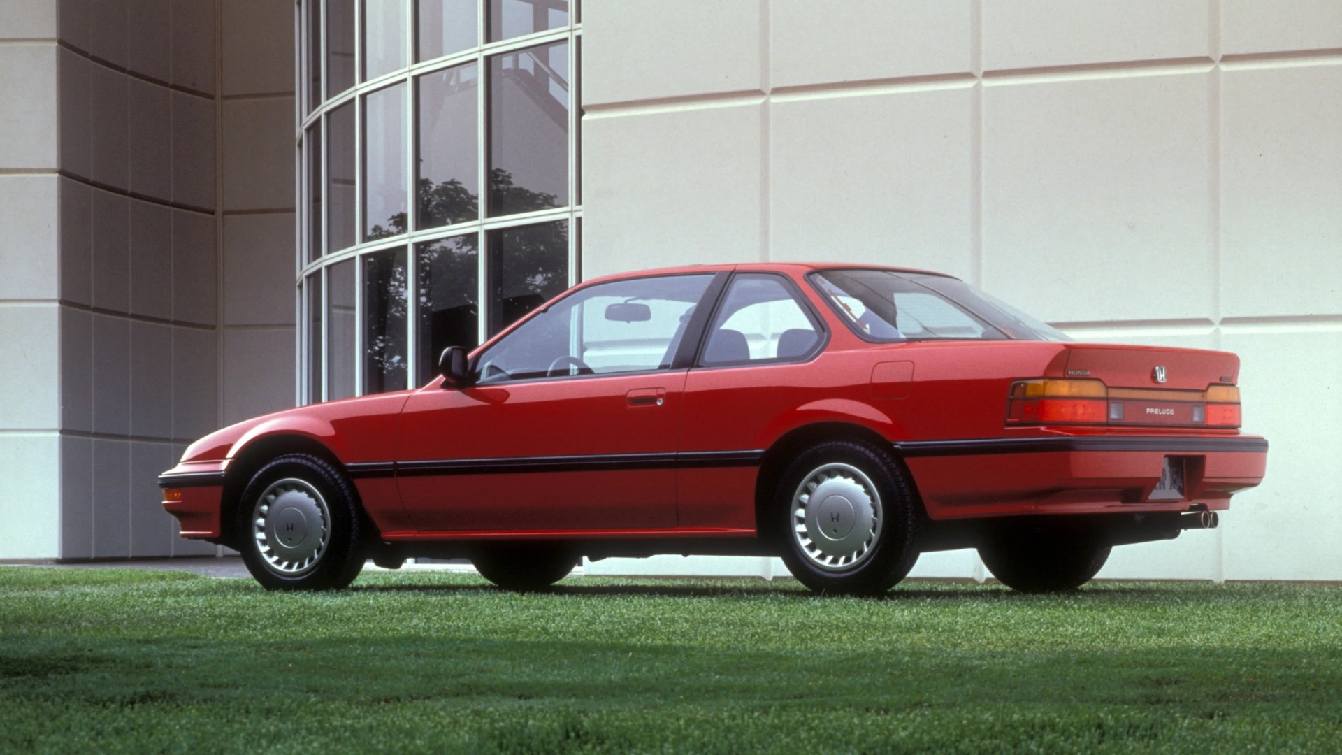 Red two-door Honda Prelude coupe parked on grass in front of a modern building with large windows.