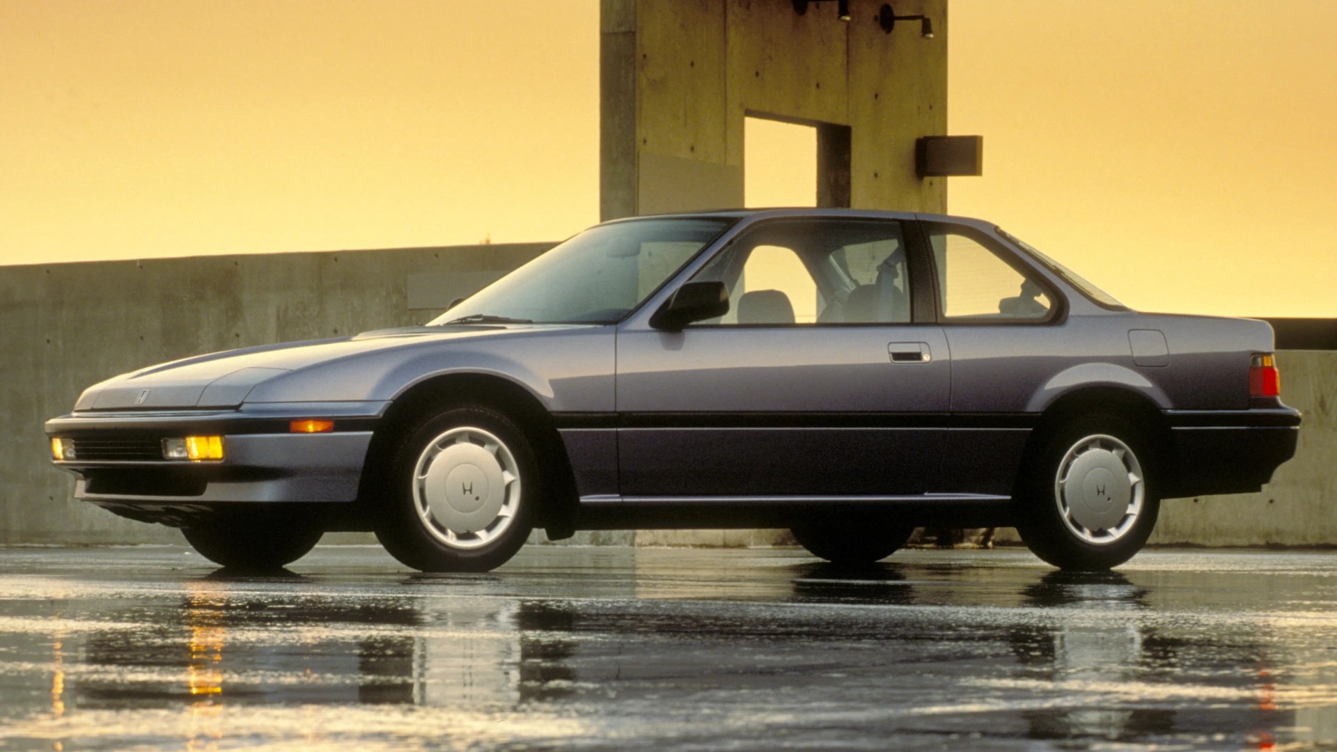 Silver two-door coupe car parked on wet pavement with concrete wall and yellow sky background.