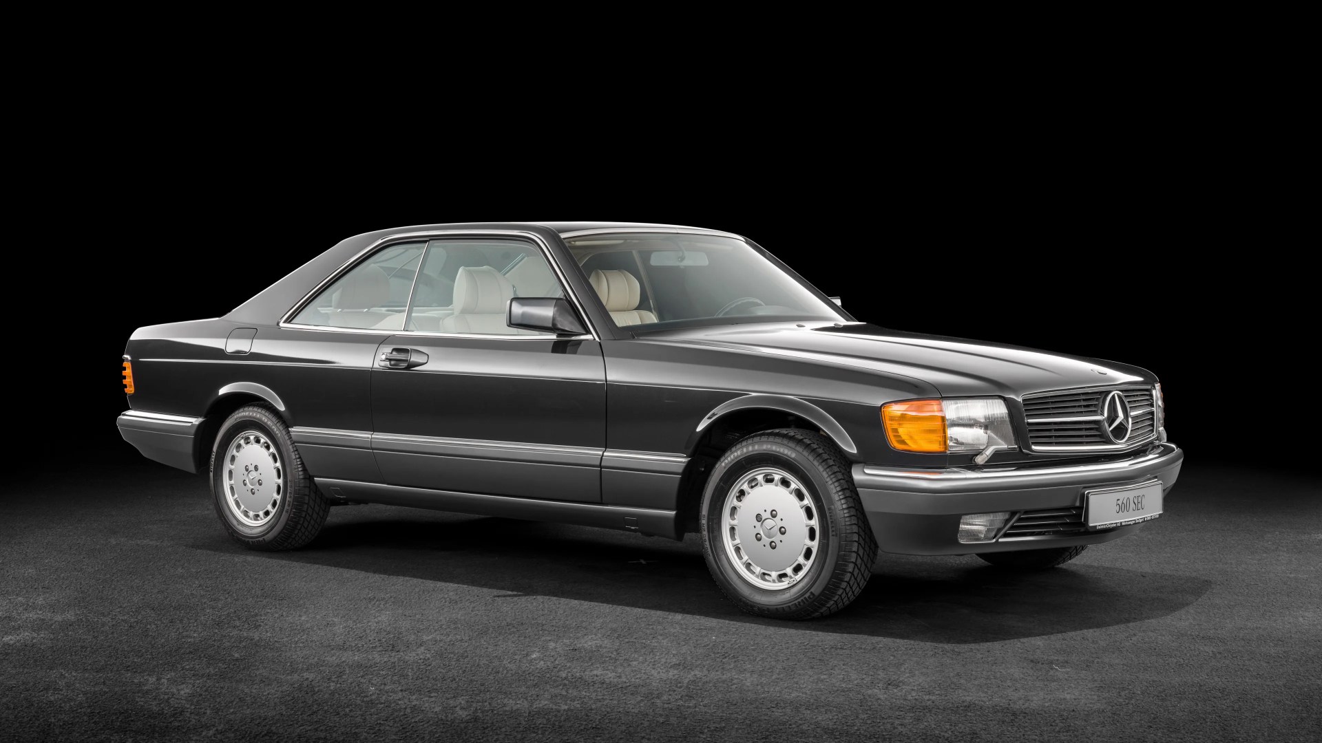 Black vintage Mercedes-Benz 560 SEC coupe with beige interior on a dark floor and black background.