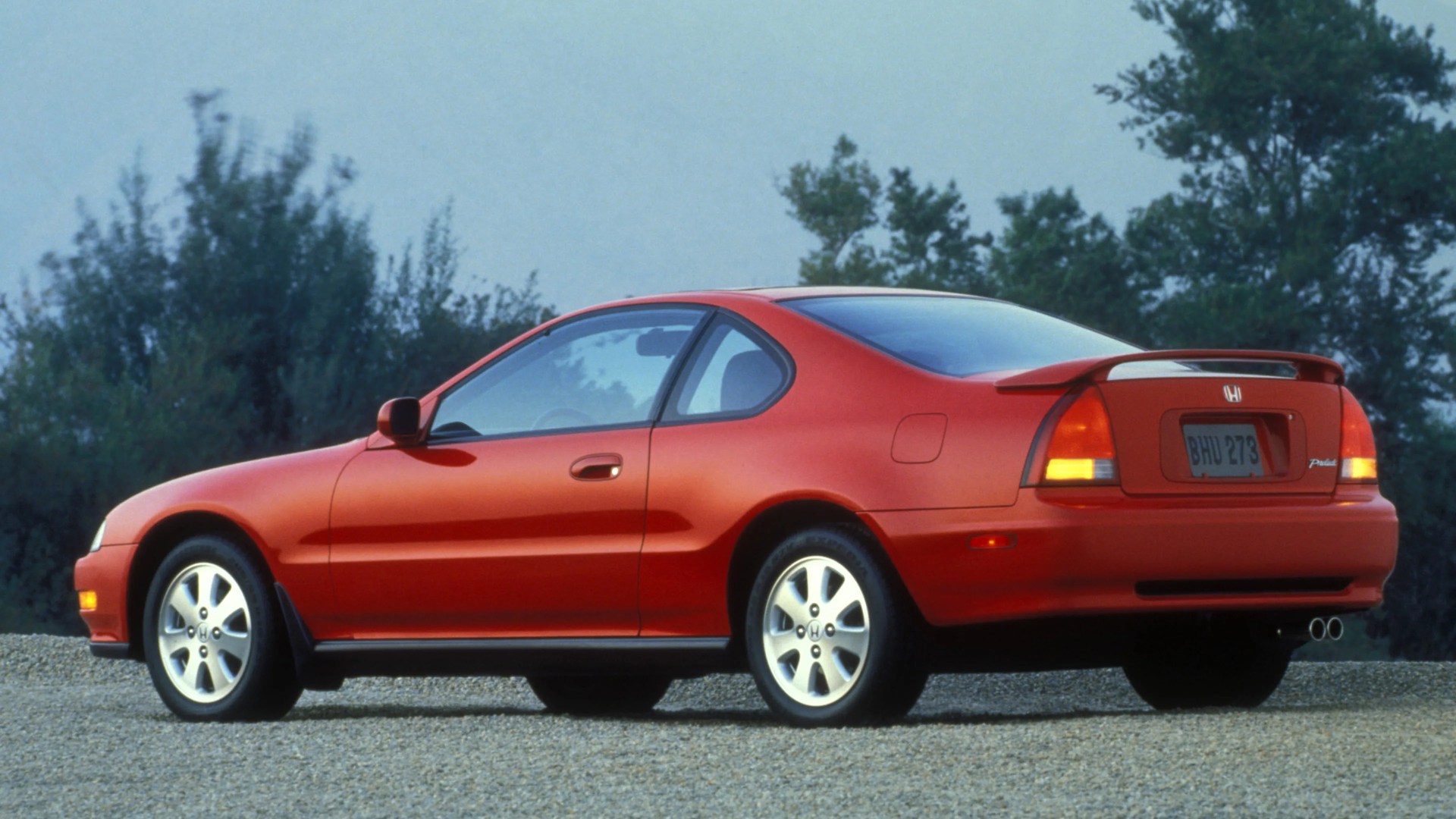Red two-door Honda Prelude coupe with rear spoiler parked on gravel with trees in the background.