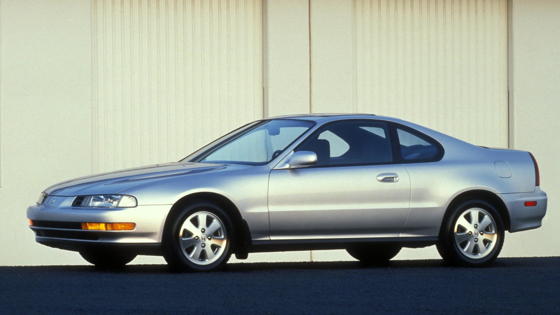 Silver two-door coupe car parked on a dark asphalt surface with a light beige wall background.