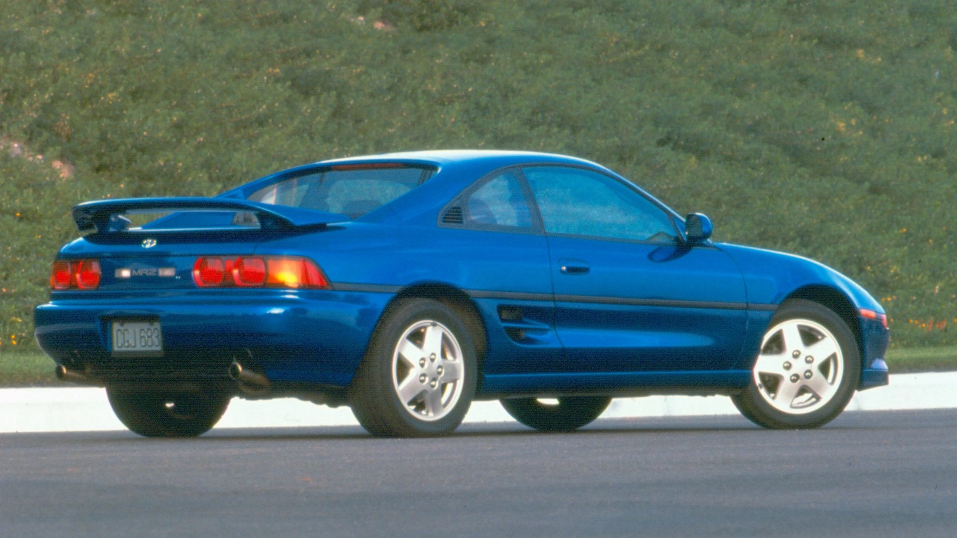Blue Toyota MR2 sports car with rear spoiler parked on a road with grass in the background.