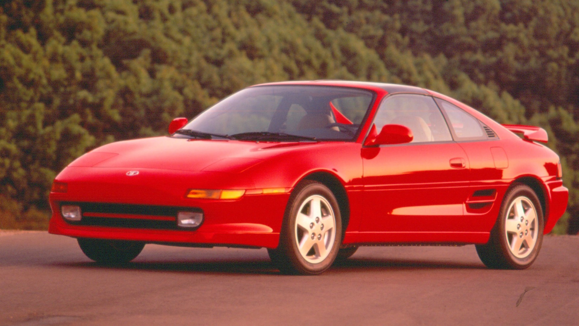 Red two-door sports coupe with pop-up headlights and rear spoiler parked on pavement.