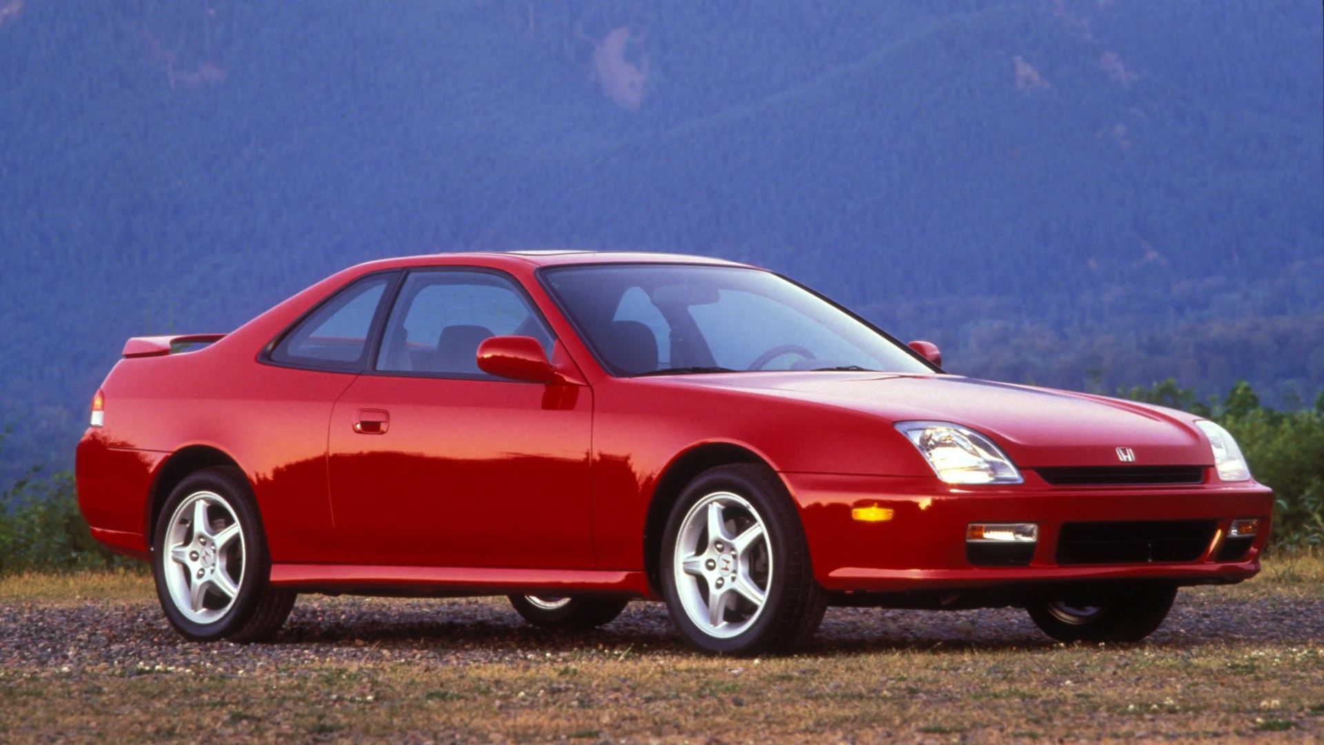 Red two-door Honda coupe with silver alloy wheels parked on gravel with mountains in the background.