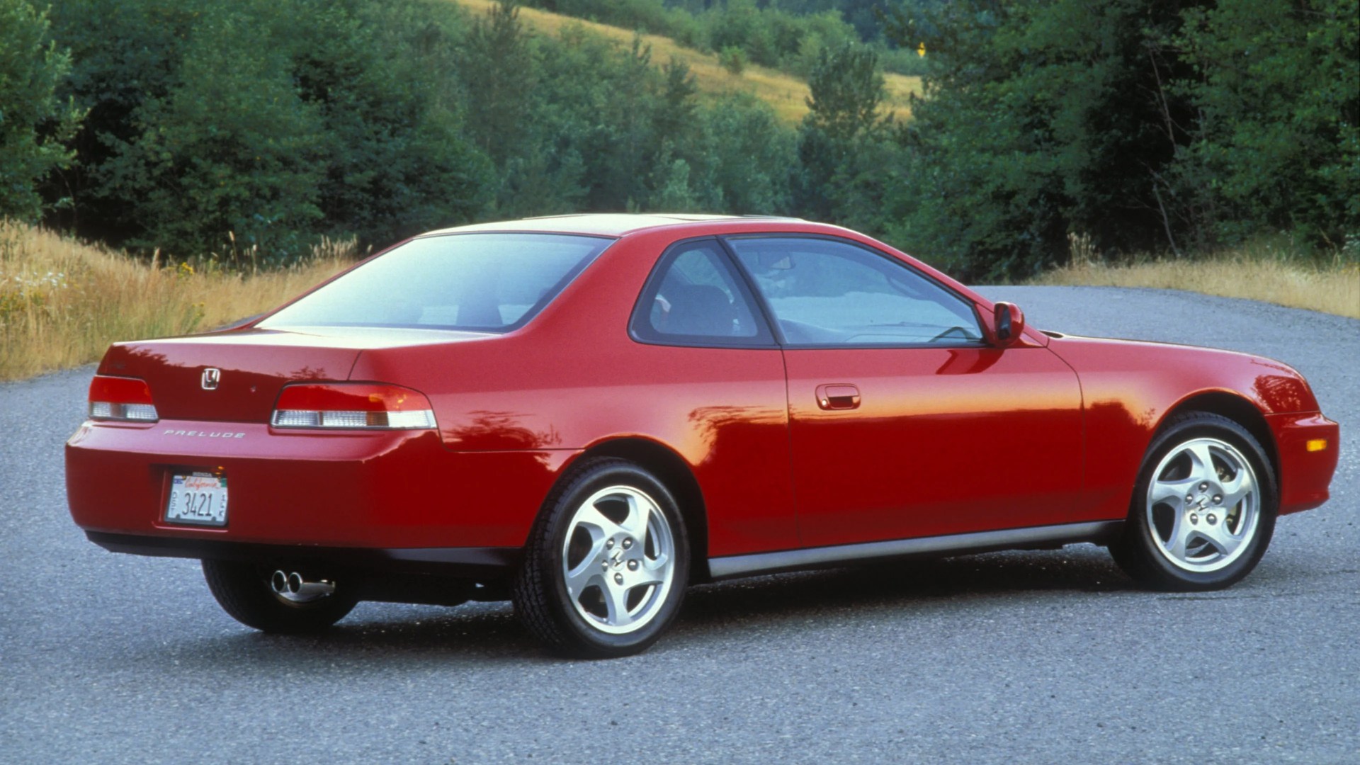 Red Honda Prelude coupe with silver alloy wheels parked on a gravel road near green trees.