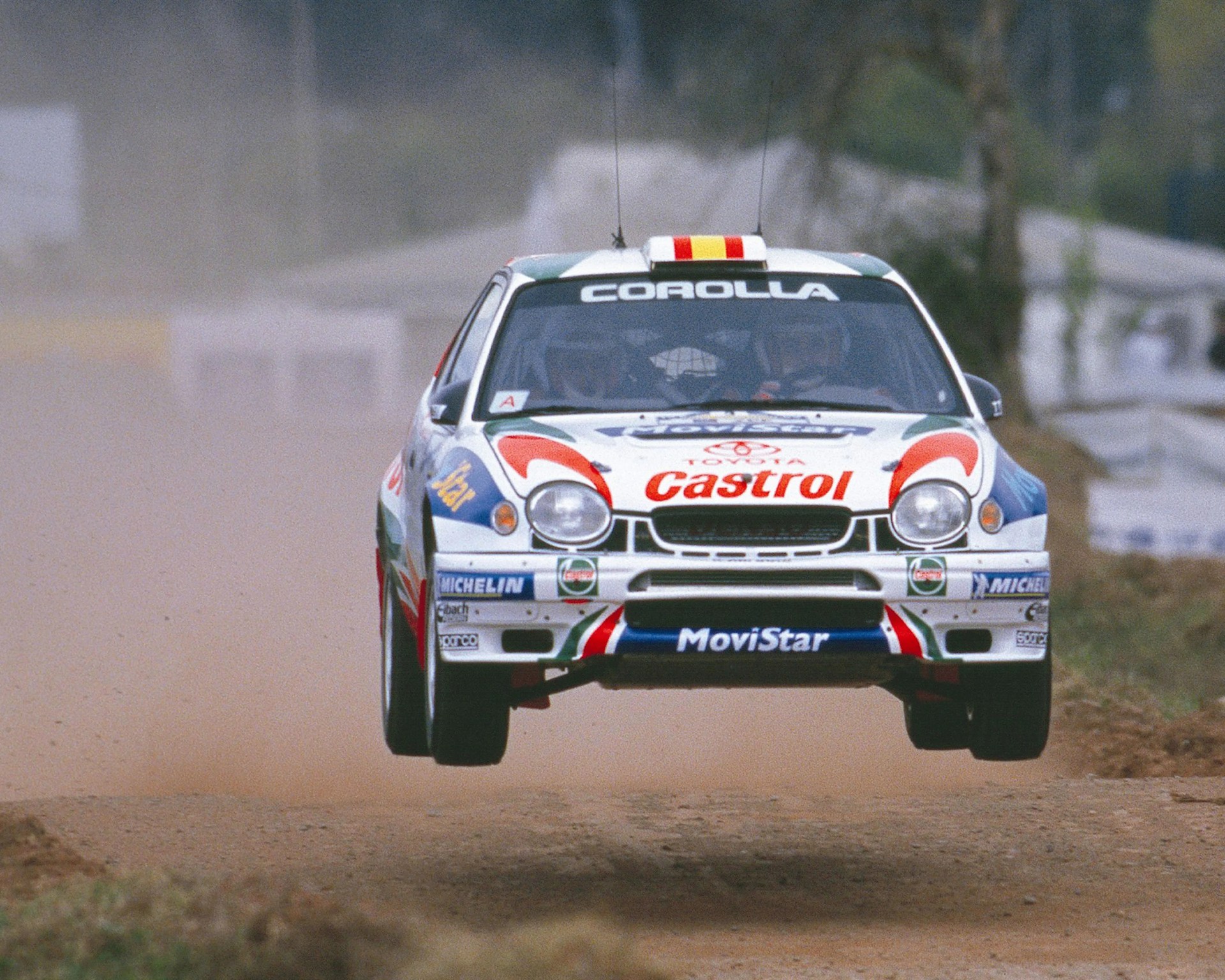 White Toyota Corolla rally car with Castrol and MoviStar logos airborne on a dirt road.