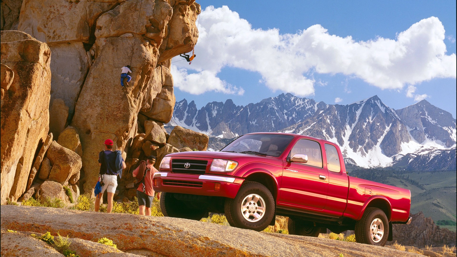 Red Toyota Tacoma pickup truck parked on rocky terrain with climbers on a large rock formation and snow-capped mountains in the background.