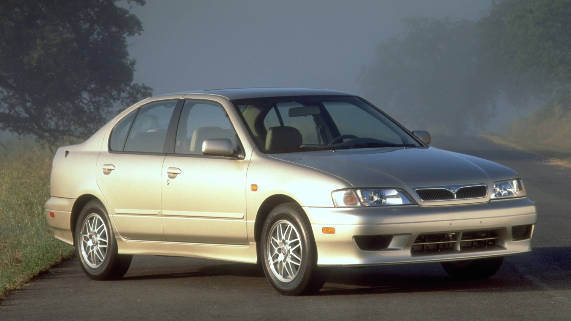 Beige four-door sedan with alloy wheels parked on a foggy road near grass and trees.