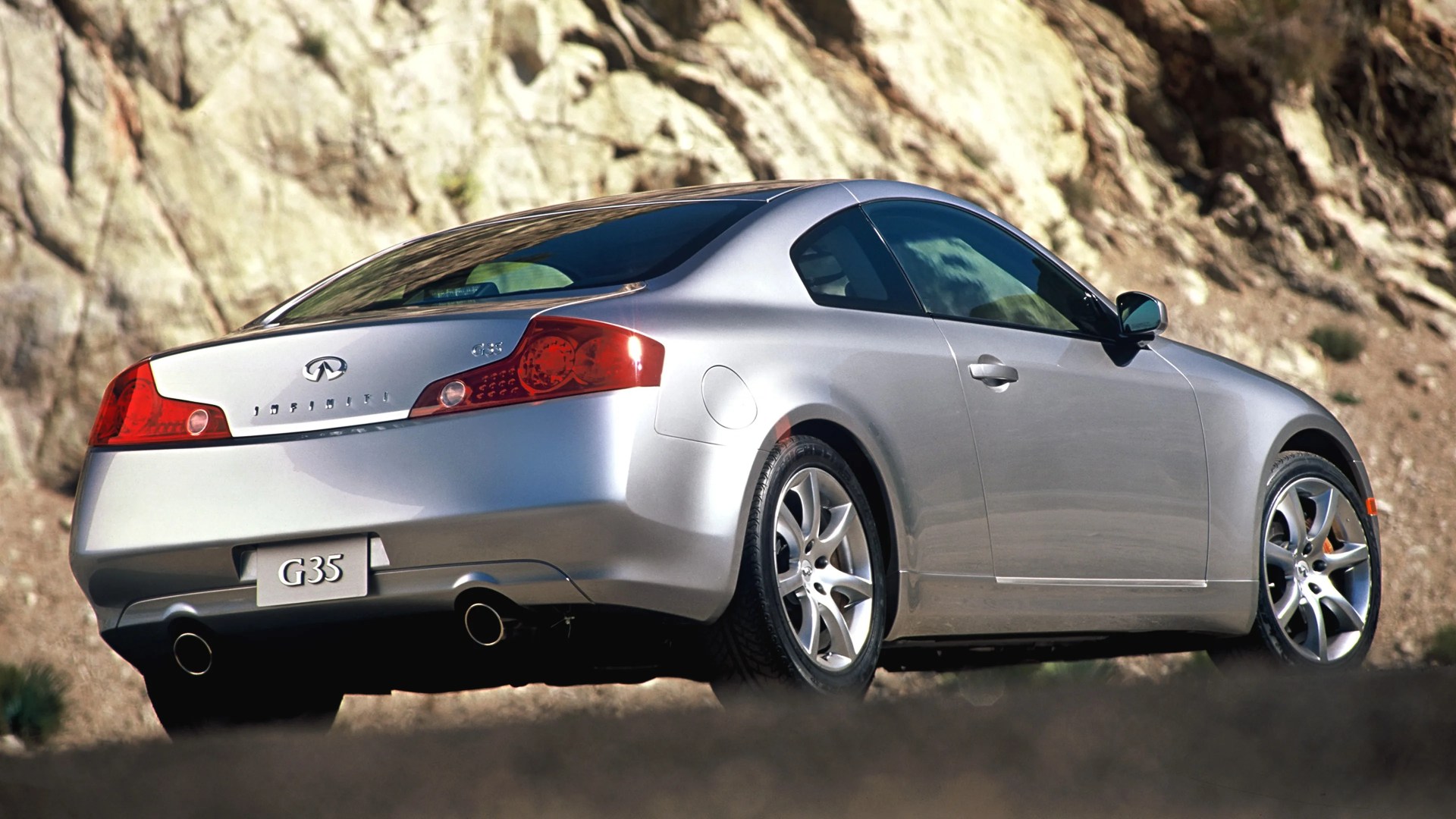 Silver Infiniti G35 coupe parked outdoors with rocky background, rear and side view.